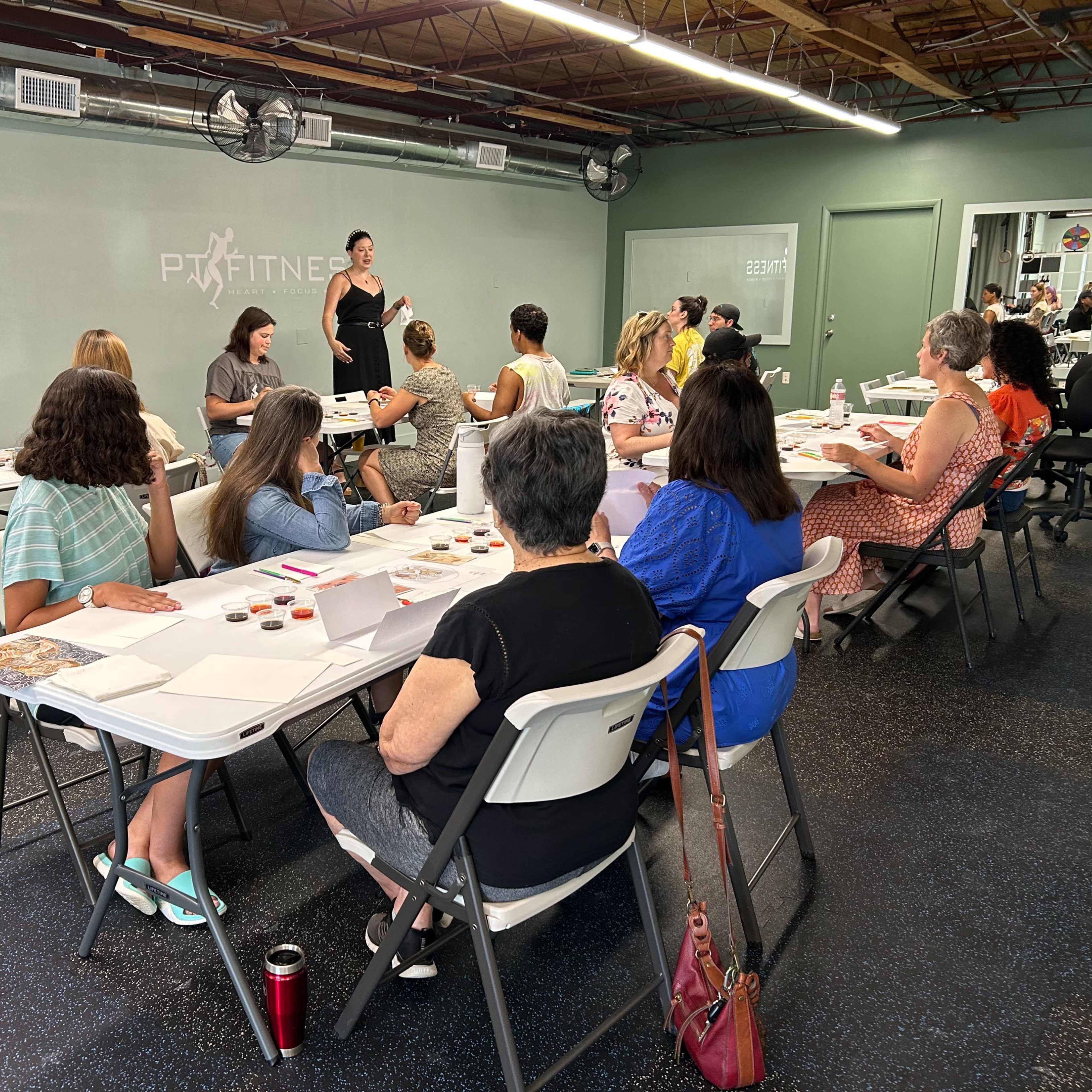 A woman stands at the front of a room filled with seated participants engaged in a workshop or class.