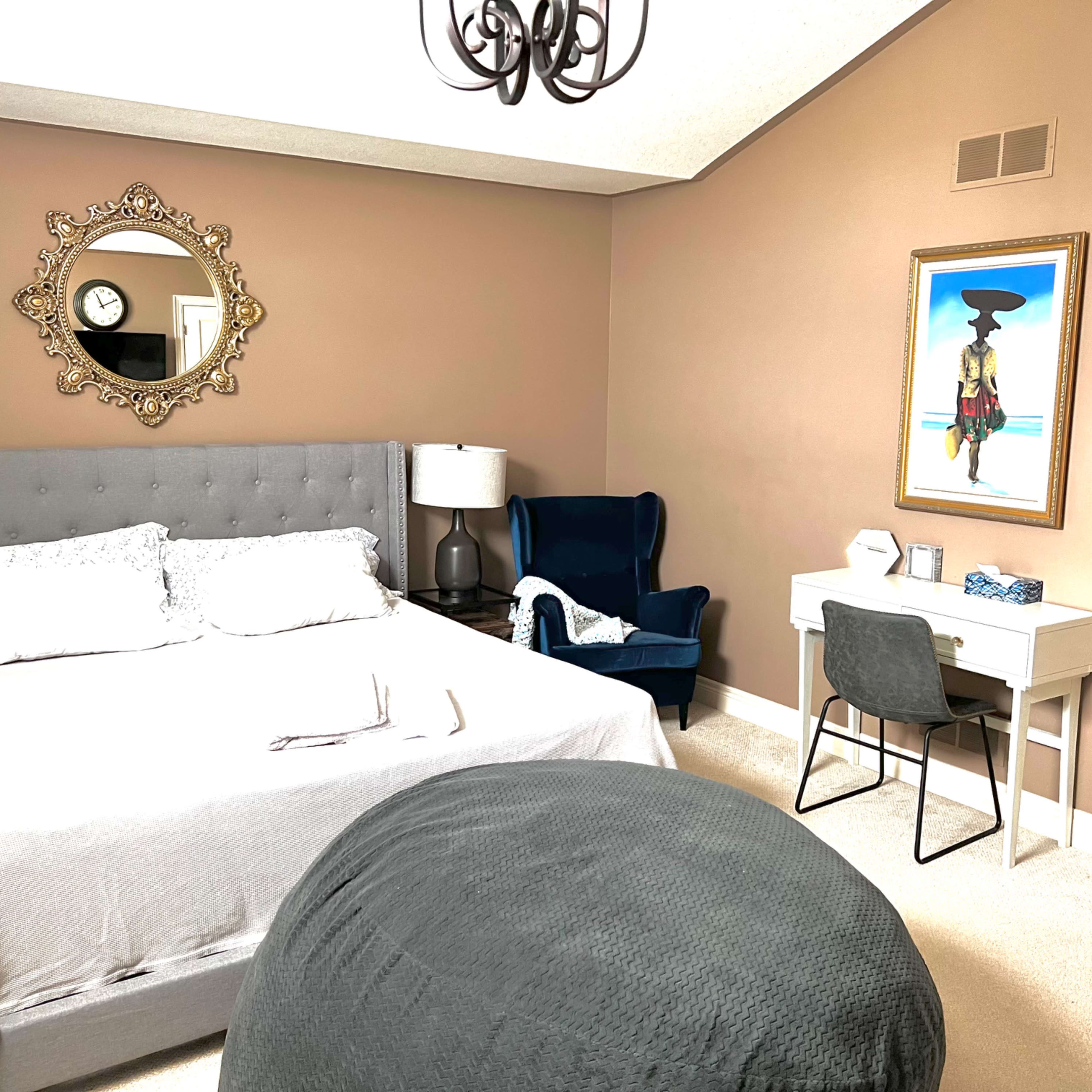 A well-decorated bedroom featuring a gray bed, a decorative mirror, a blue armchair, and a small desk with a chair.