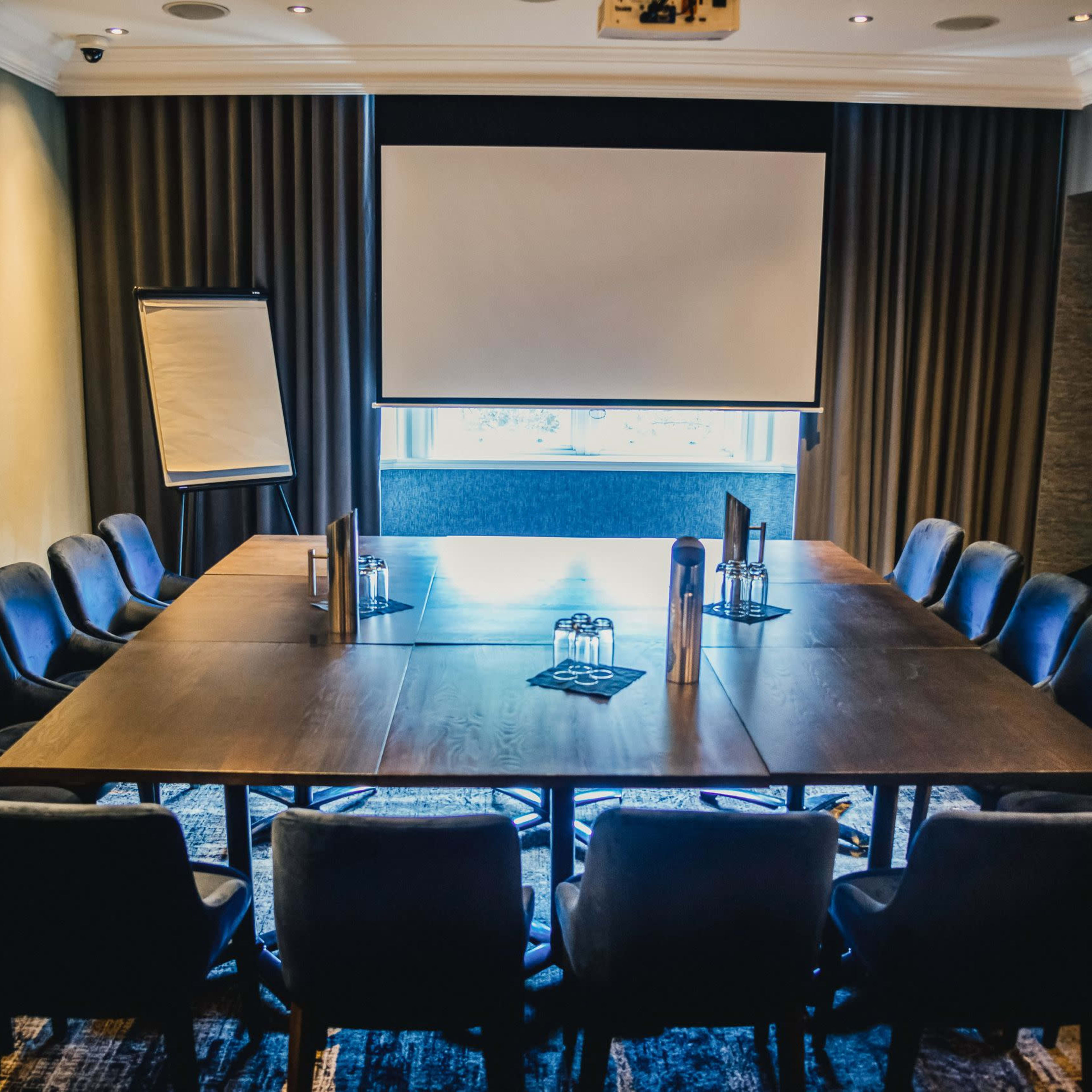A conference room features a large rectangular table surrounded by chairs, with a screen and a flip chart at one end.