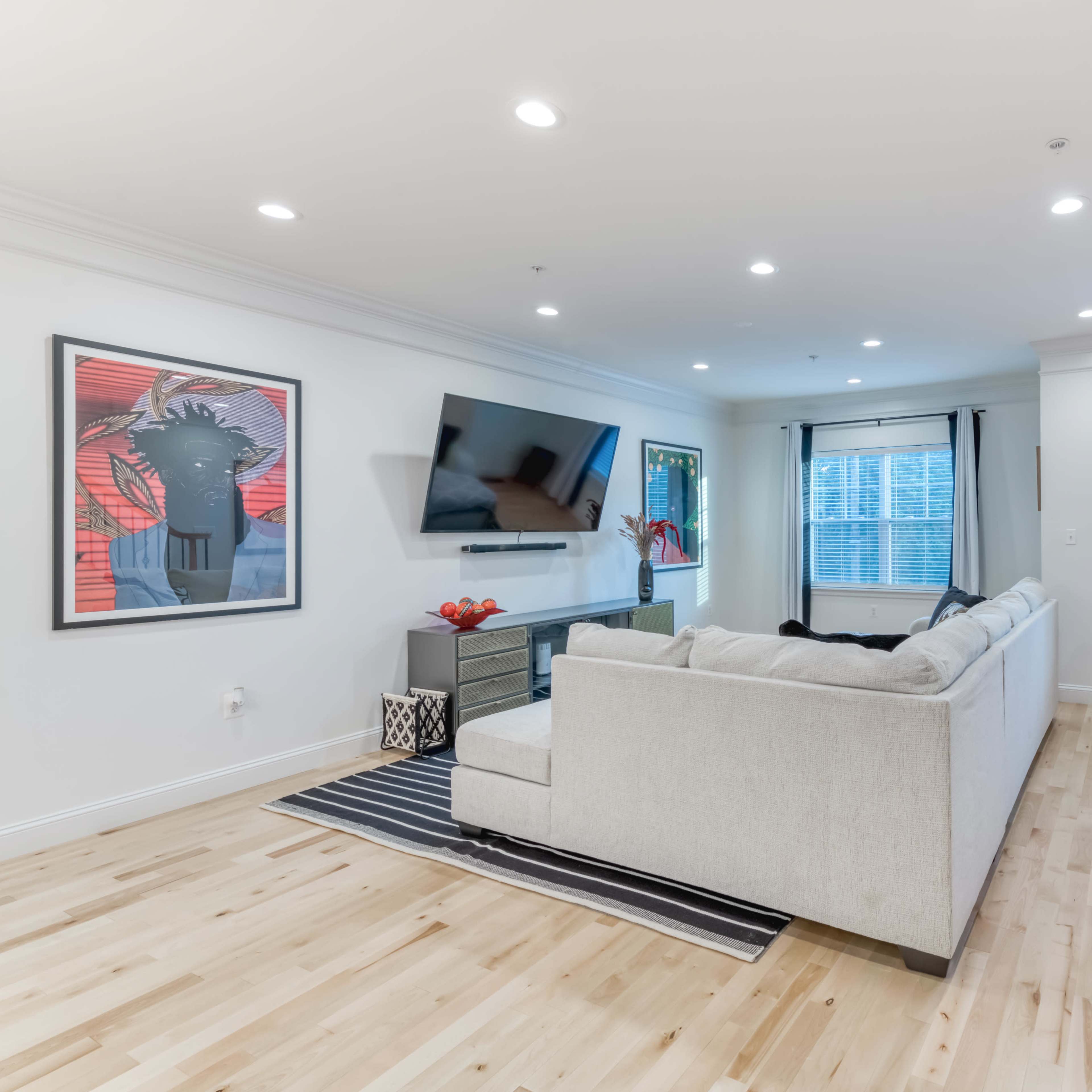 A spacious living room features a sectional sofa, a mounted TV, hardwood flooring, and large windows with curtains.