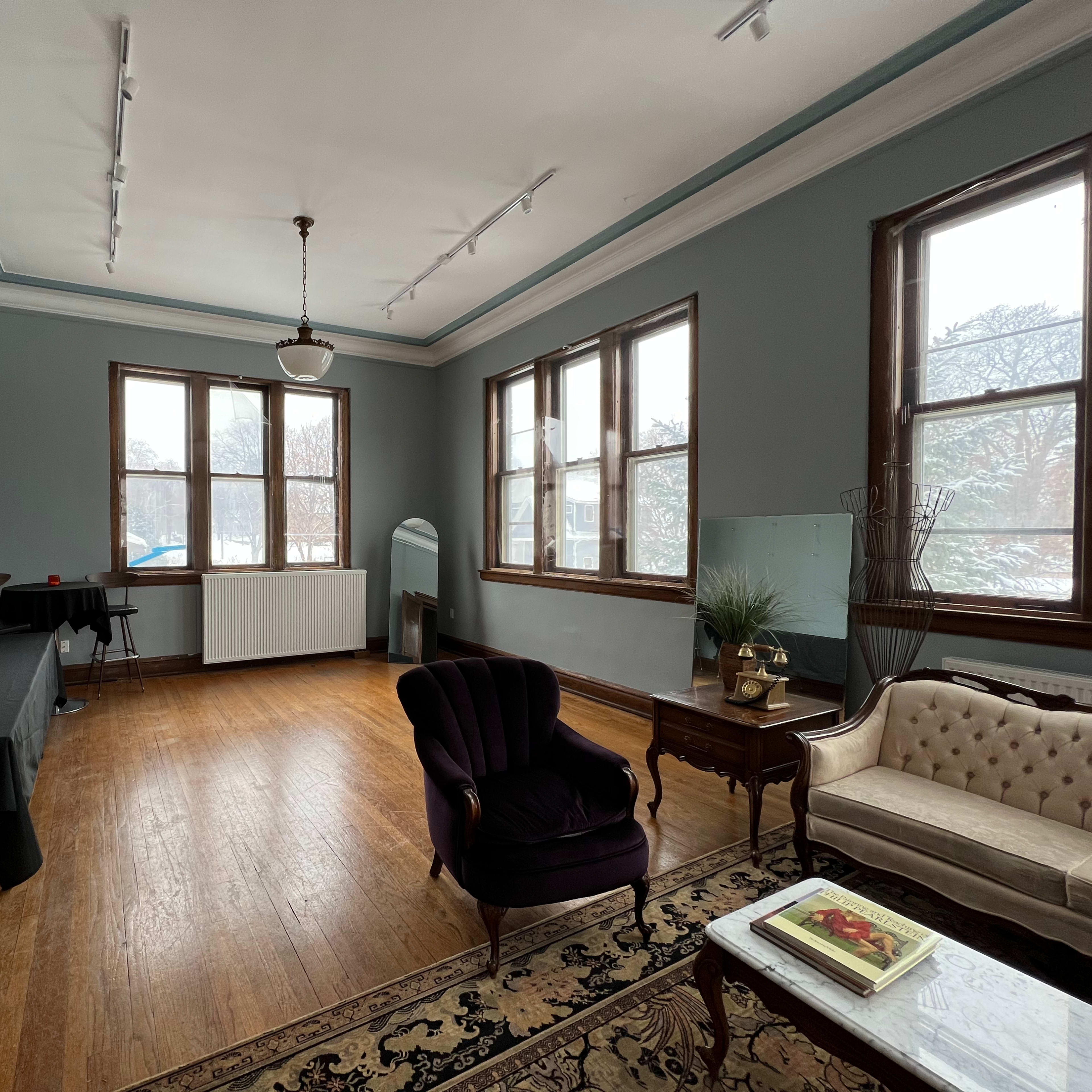 A spacious, empty room with large windows, hardwood floors, and a mixture of furniture including a velvet armchair and a couch.