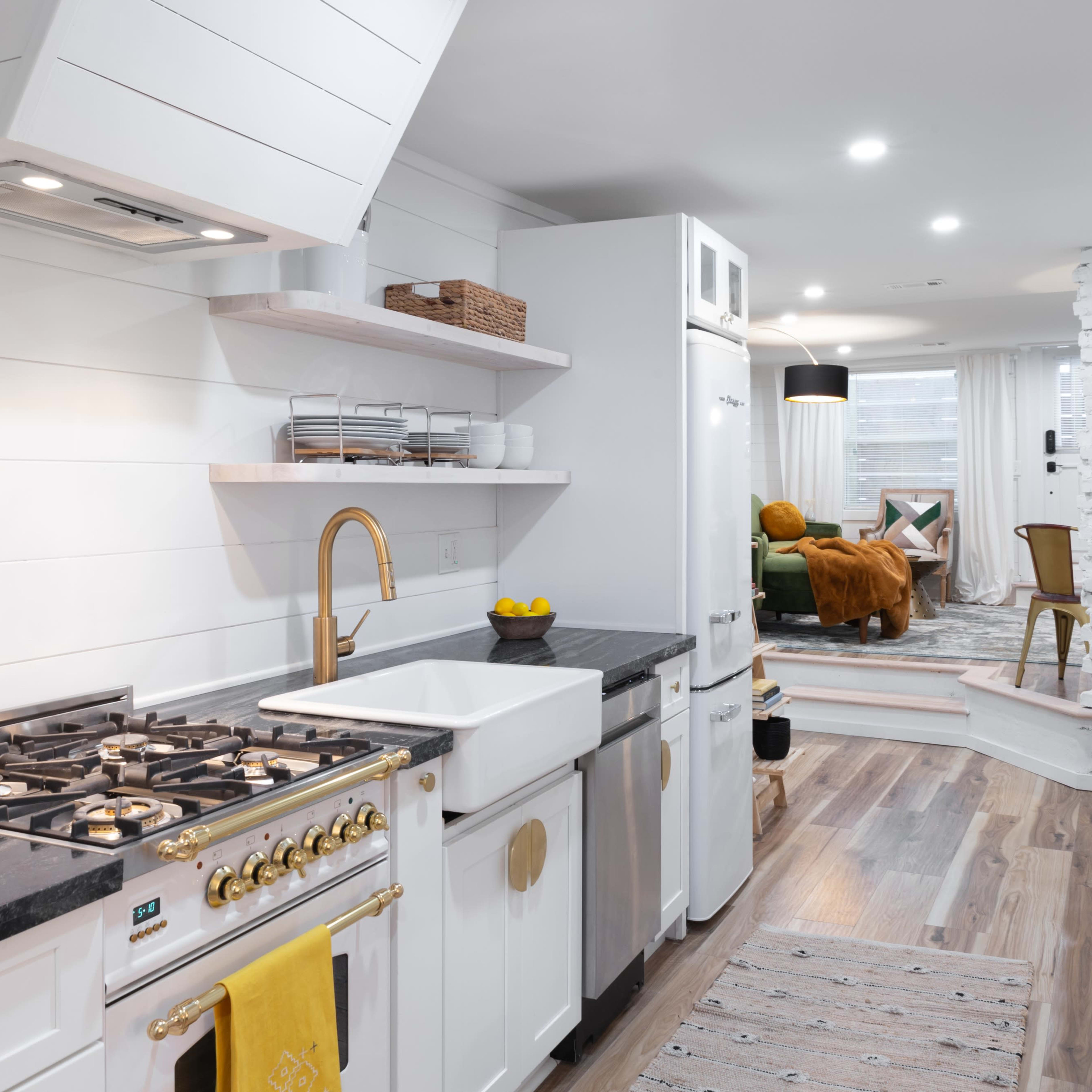 A modern kitchen featuring a gas range, farmhouse sink, white cabinetry, and open shelving, with a living area visible in the background.