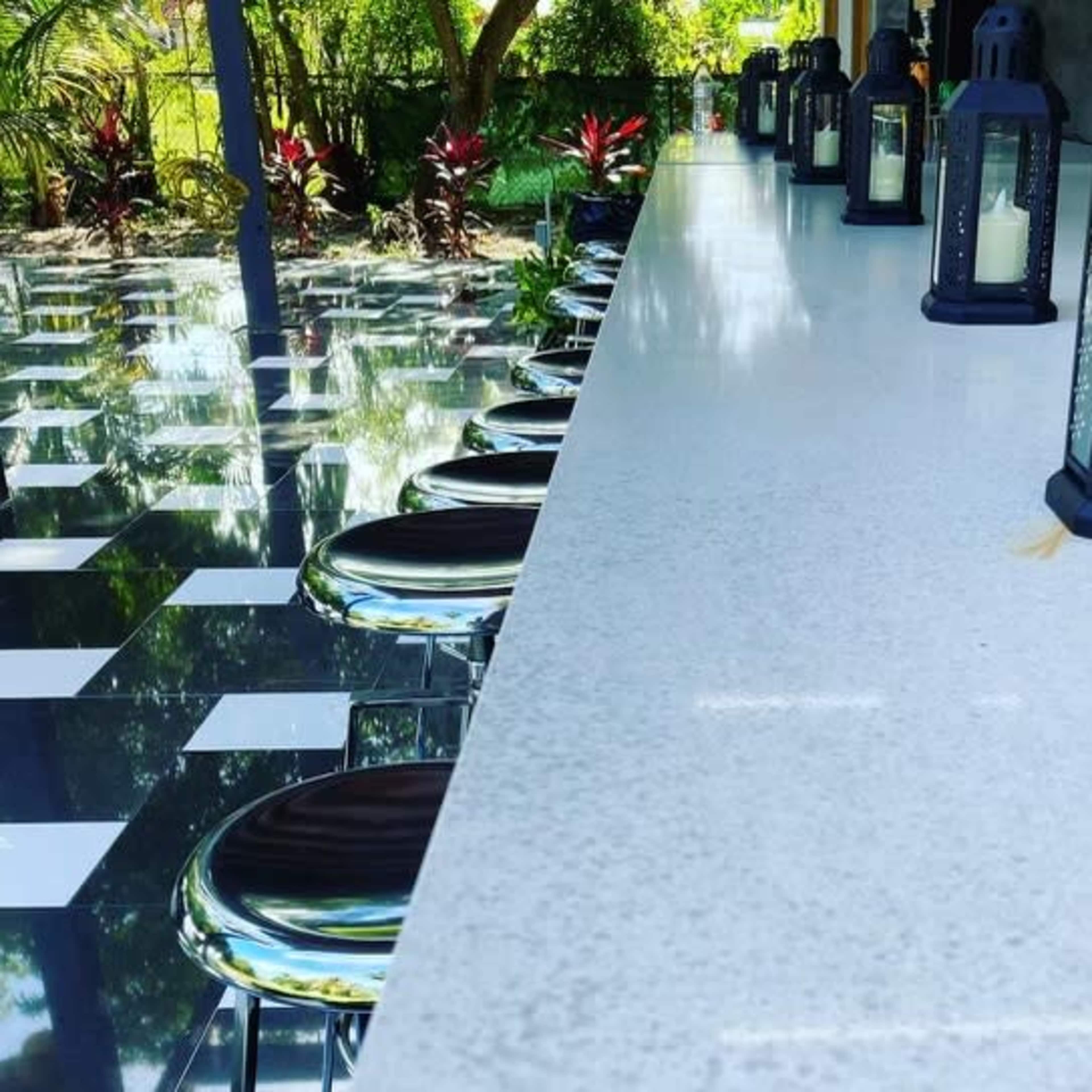 A long, white countertop with black lanterns is lined with metallic stools, set in a garden with a checkerboard floor and greenery.