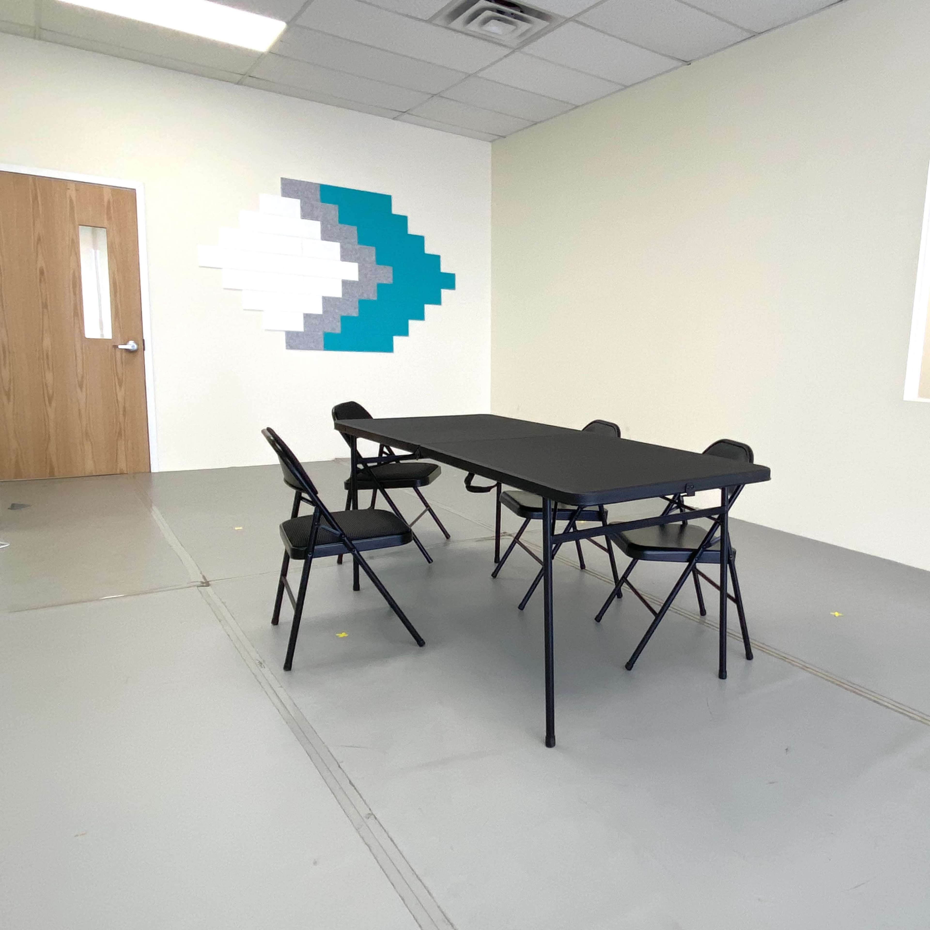 A rectangular table with four folding chairs is positioned in a room that features a wall decoration of teal and white geometric shapes.