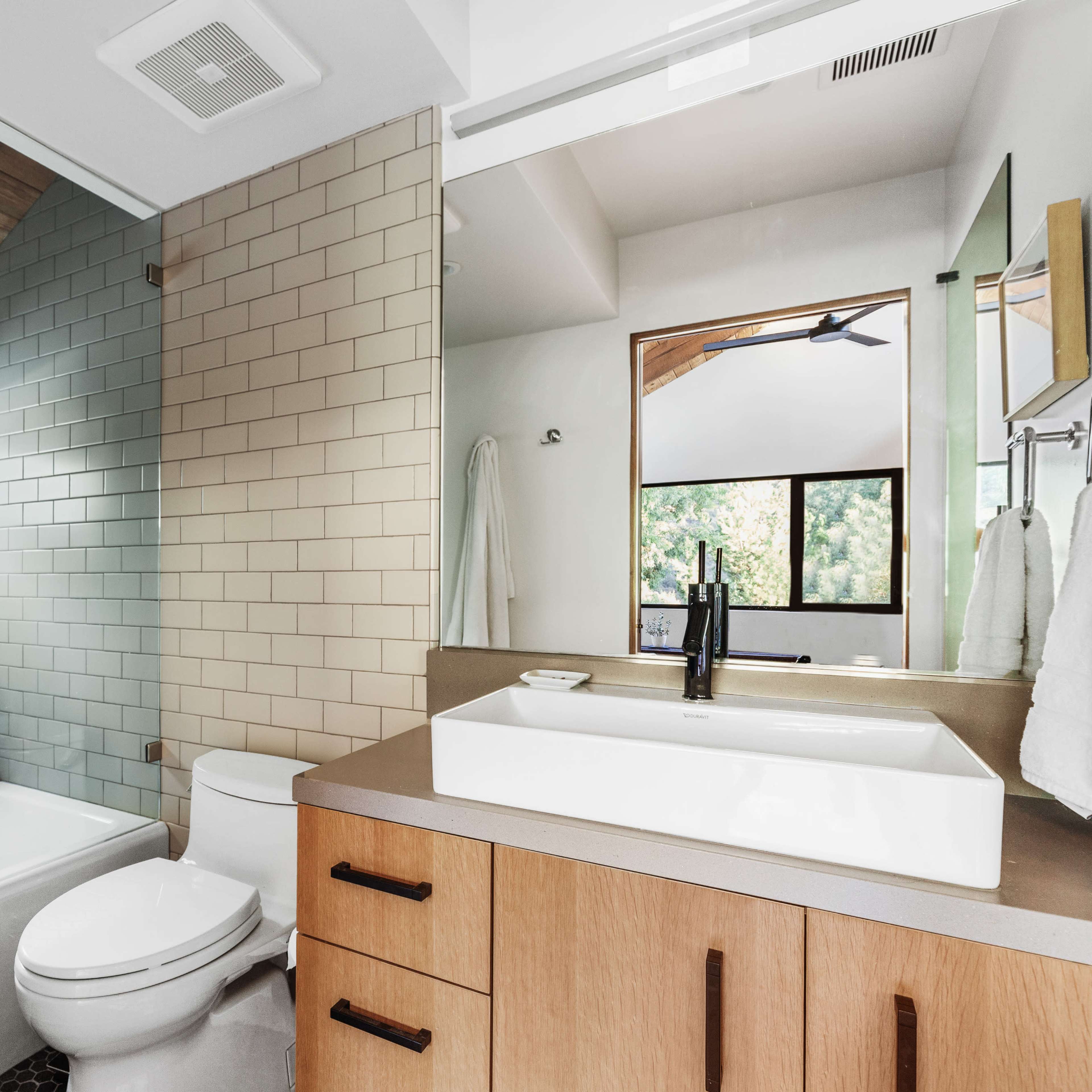 The bathroom features a glass shower enclosure, a freestanding bathtub, a modern sink with wood cabinetry, and a large window allowing natural light.