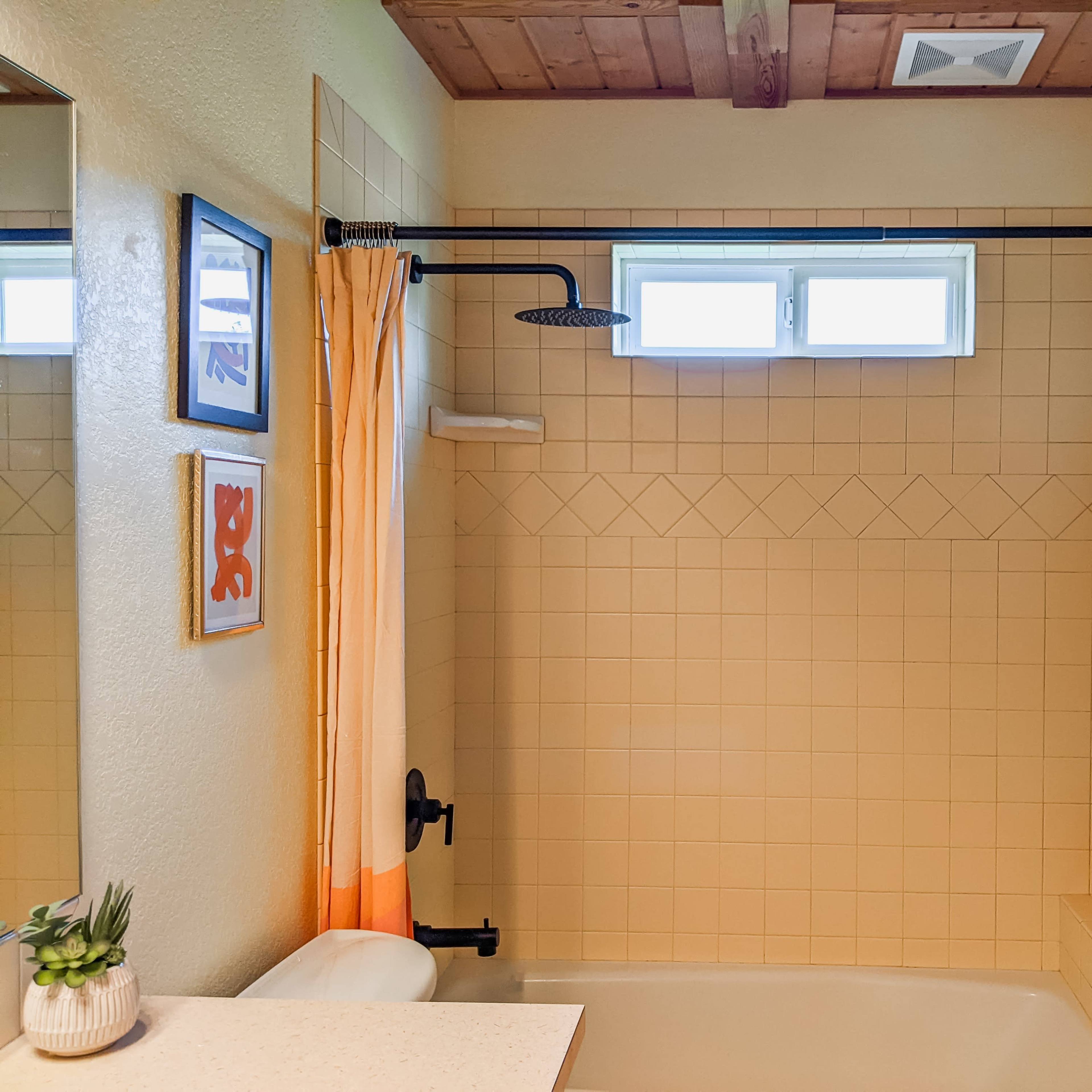 A bathroom with a tiled shower area, featuring a showerhead, a window, and a curtain in orange tones.