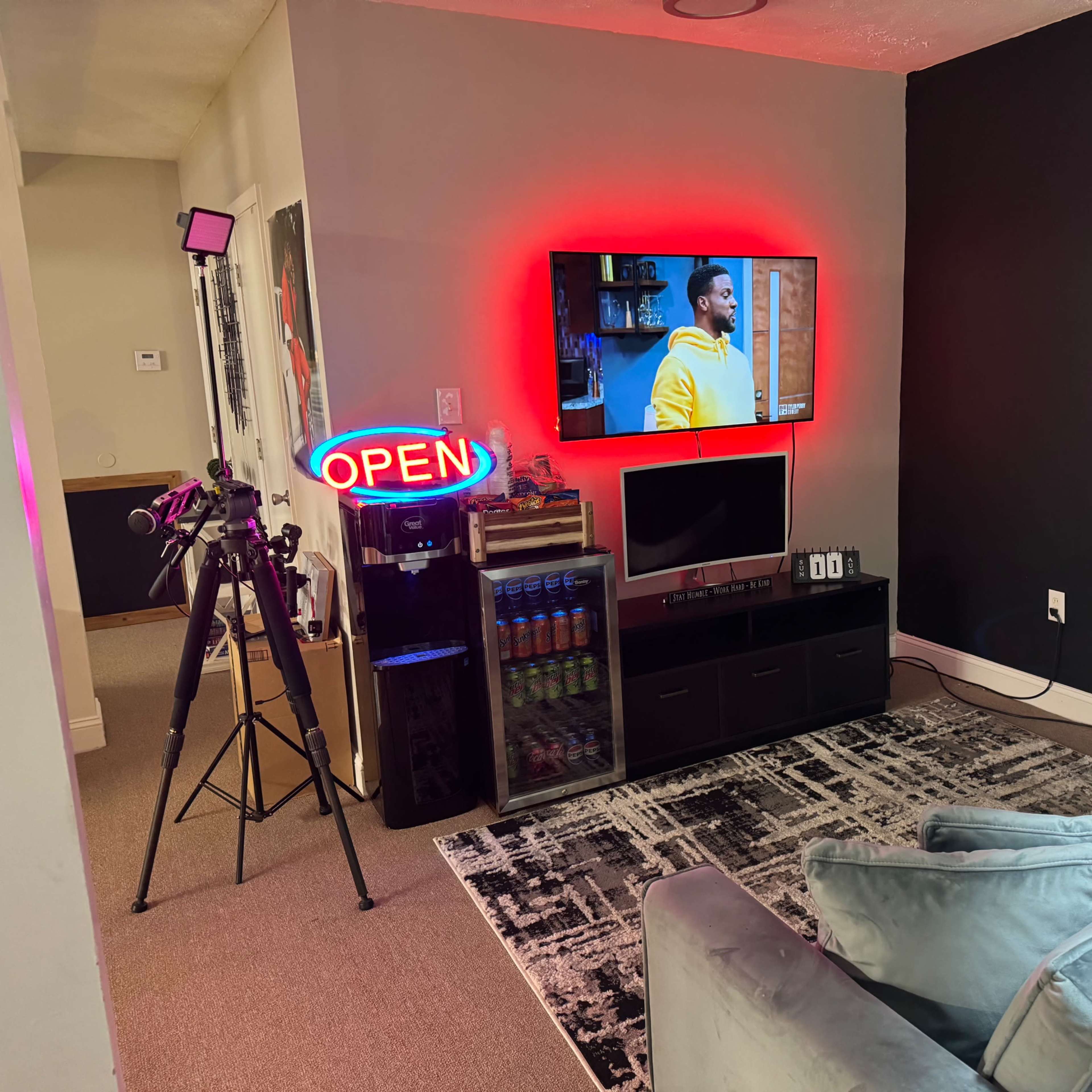 A living room setup with a large TV displaying a show, a neon "OPEN" sign, a camera on a tripod, and a shelf with various items, all against a dark wall.