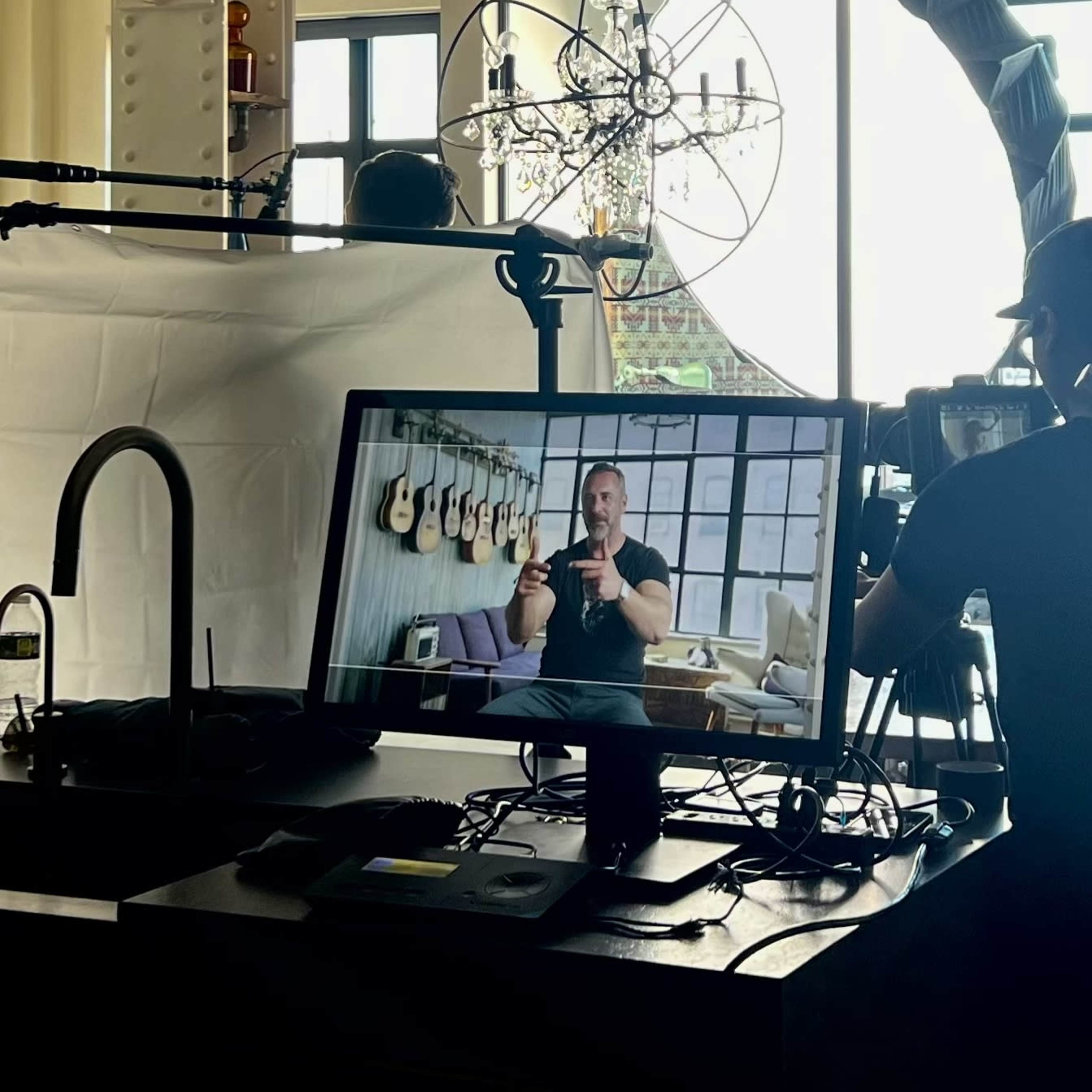 A video production setup is visible, featuring a monitor displaying a person speaking, with cameras and lighting equipment arranged around a large window.