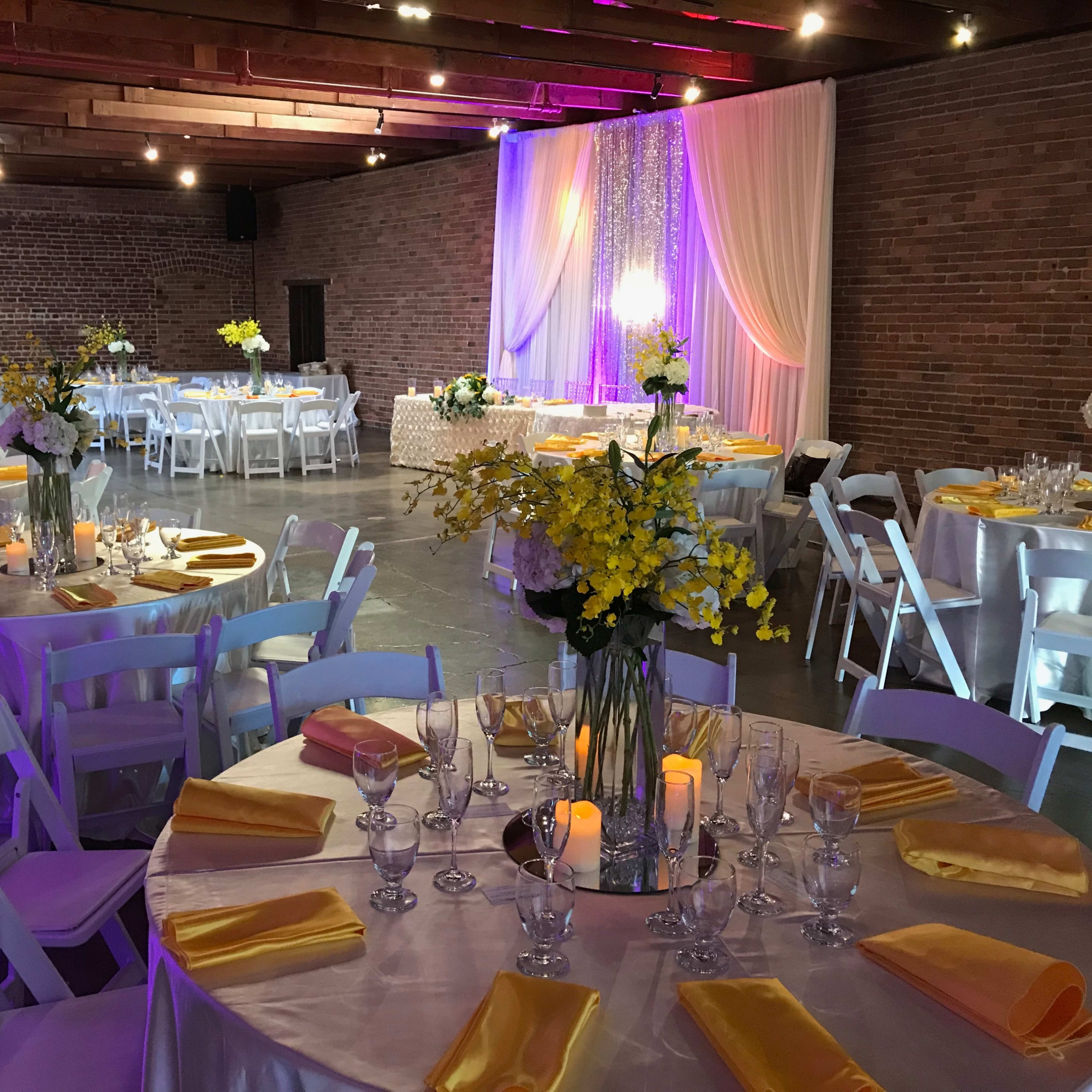The venue features elegantly set round tables with yellow napkins and floral centerpieces, under soft lighting and draped fabric against a brick wall.