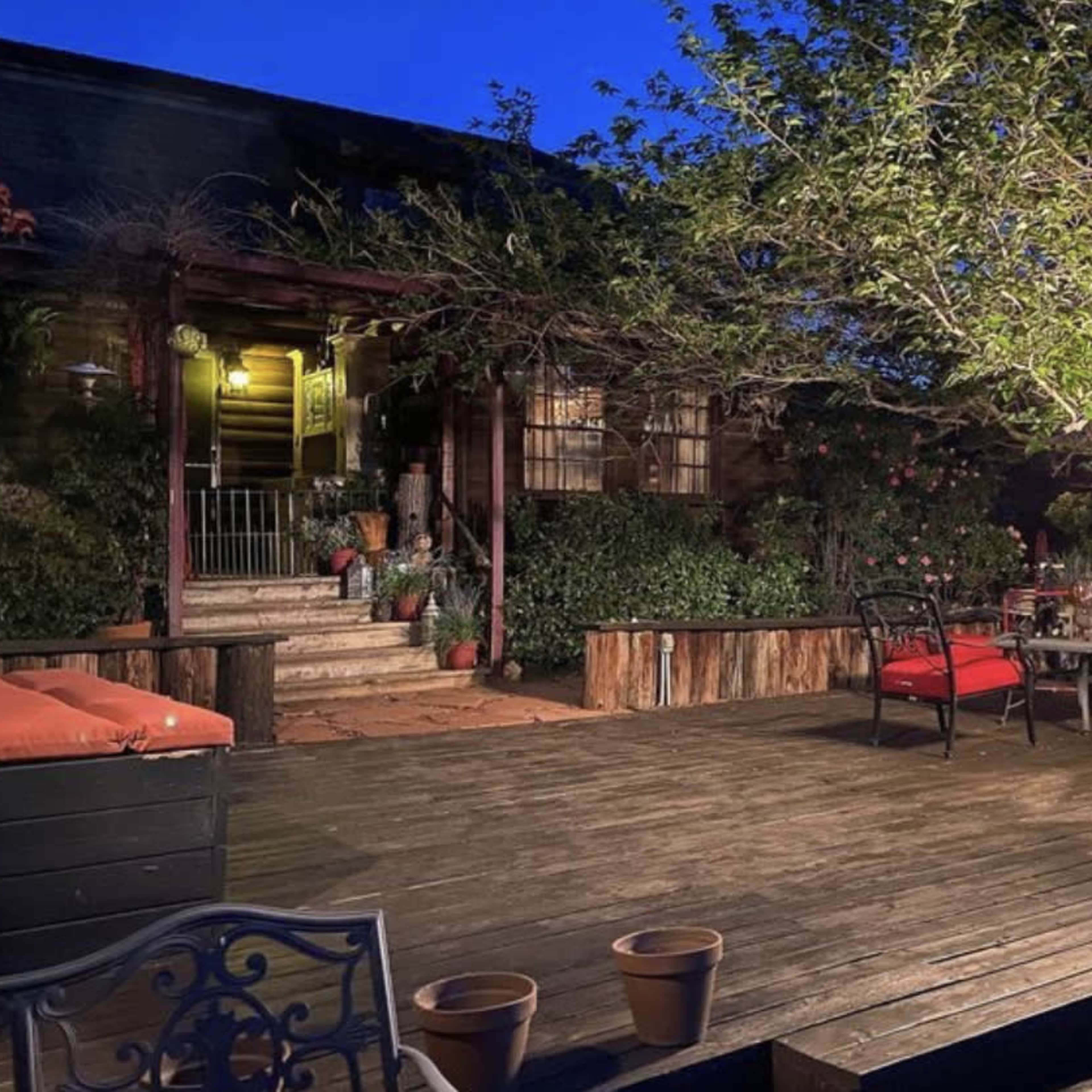 A wooden deck area at dusk, featuring outdoor seating with red cushions, a lantern, and surrounding greenery.