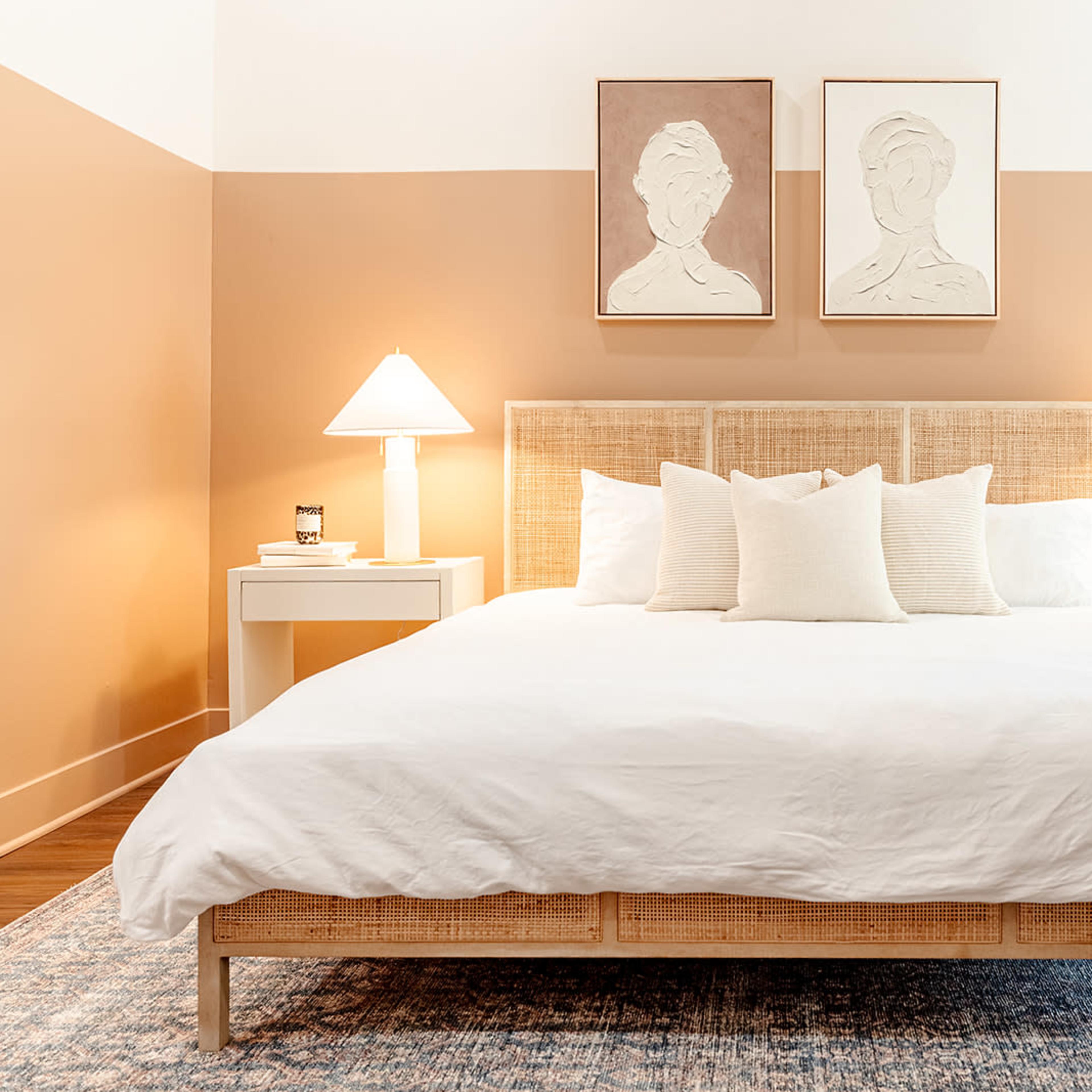 A bright bedroom featuring a large white bed with multiple pillows, flanked by two white bedside tables with lamps, and decorated with minimalist artwork on the walls.