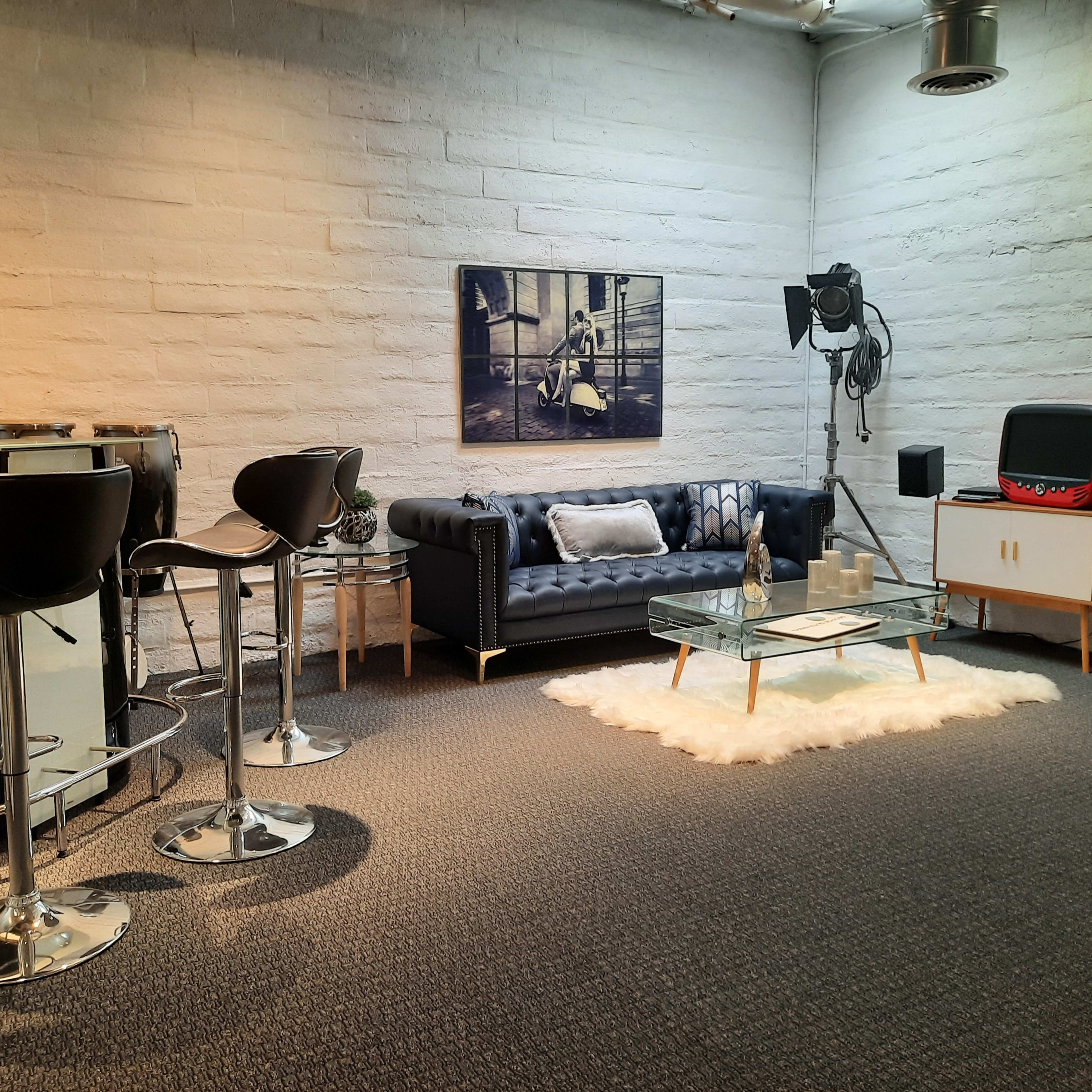A modern lounge area featuring a black leather sofa, a glass coffee table on a white rug, barstools, and retro television.