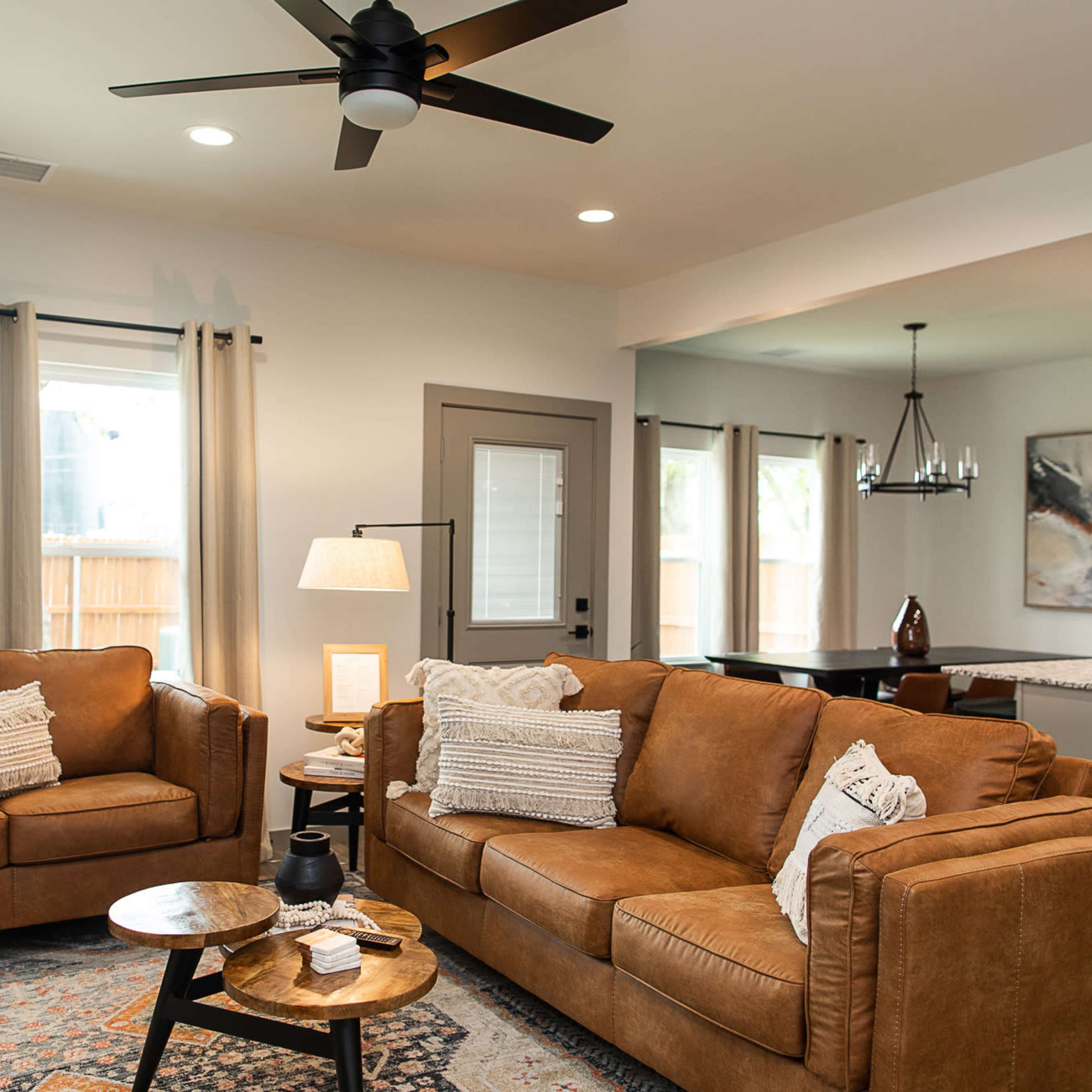 The living room features two brown leather sofas, a wooden side table, and light-colored curtains, with a dining area visible in the background.