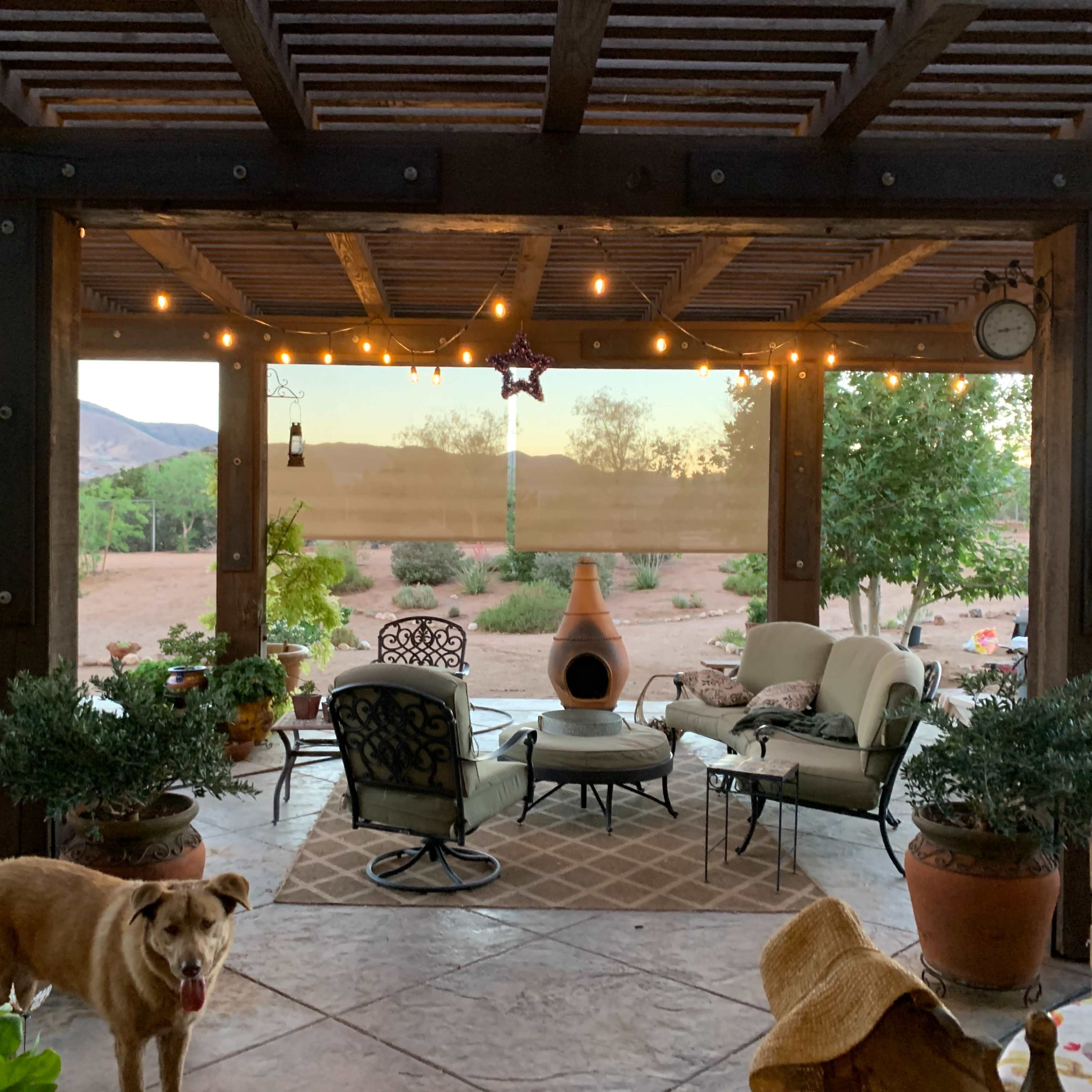 A covered patio area features chairs and a table surrounded by potted plants, with a view of a desert landscape and string lights overhead.