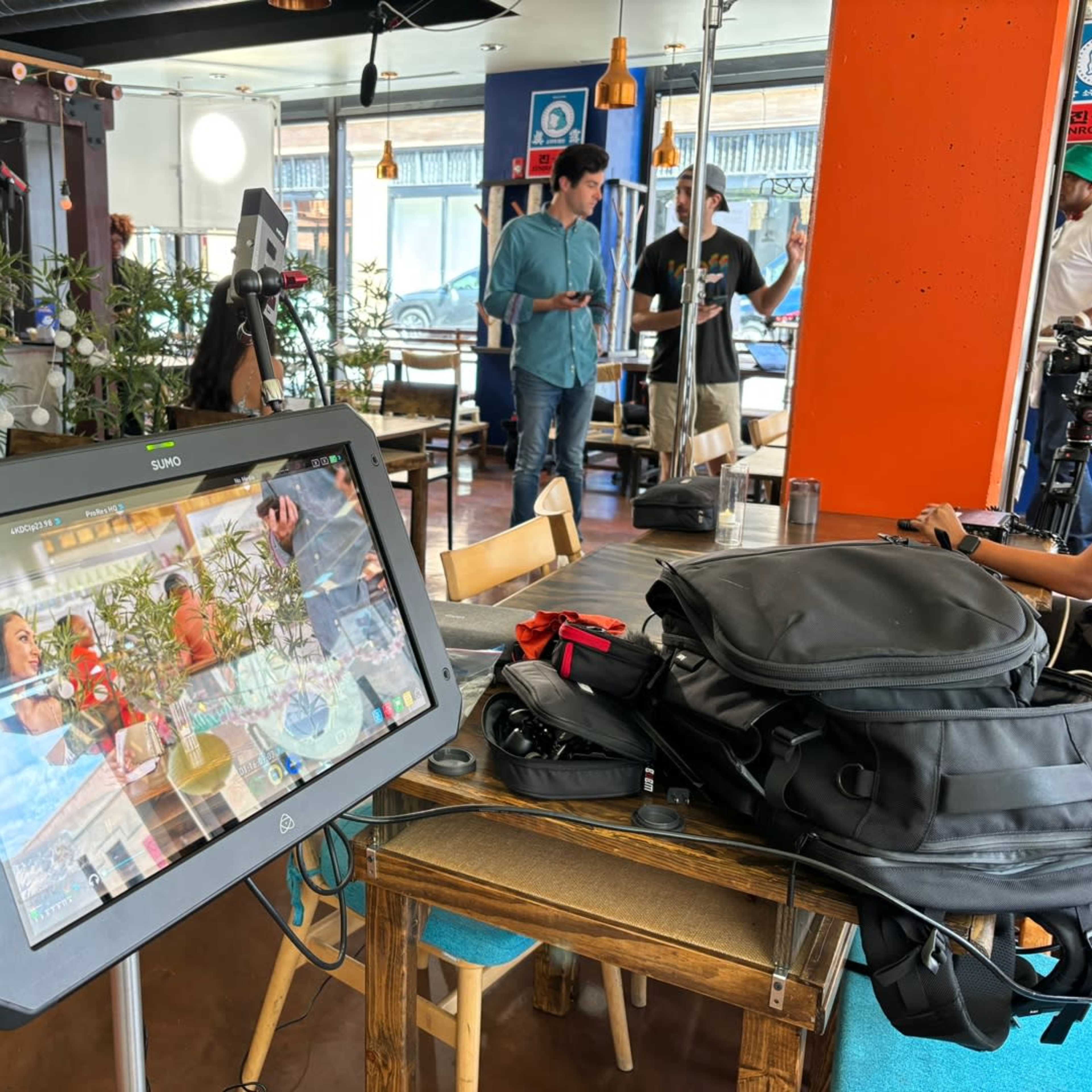 A digital monitor displays a video feed while crew members set up equipment in a café setting.