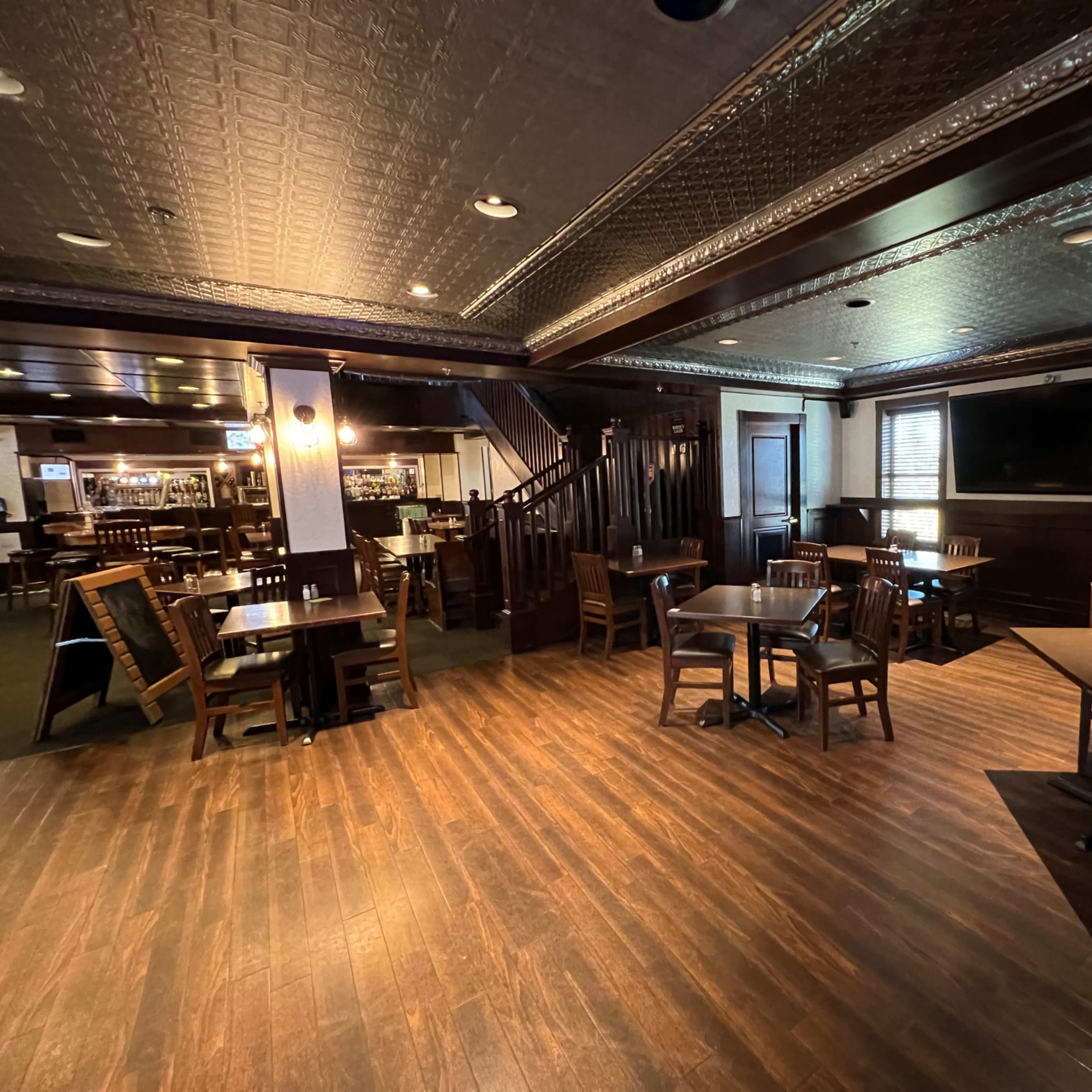 An interior view of a dimly lit restaurant with wooden tables and chairs arranged on a hardwood floor, featuring a staircase and a bar in the background.