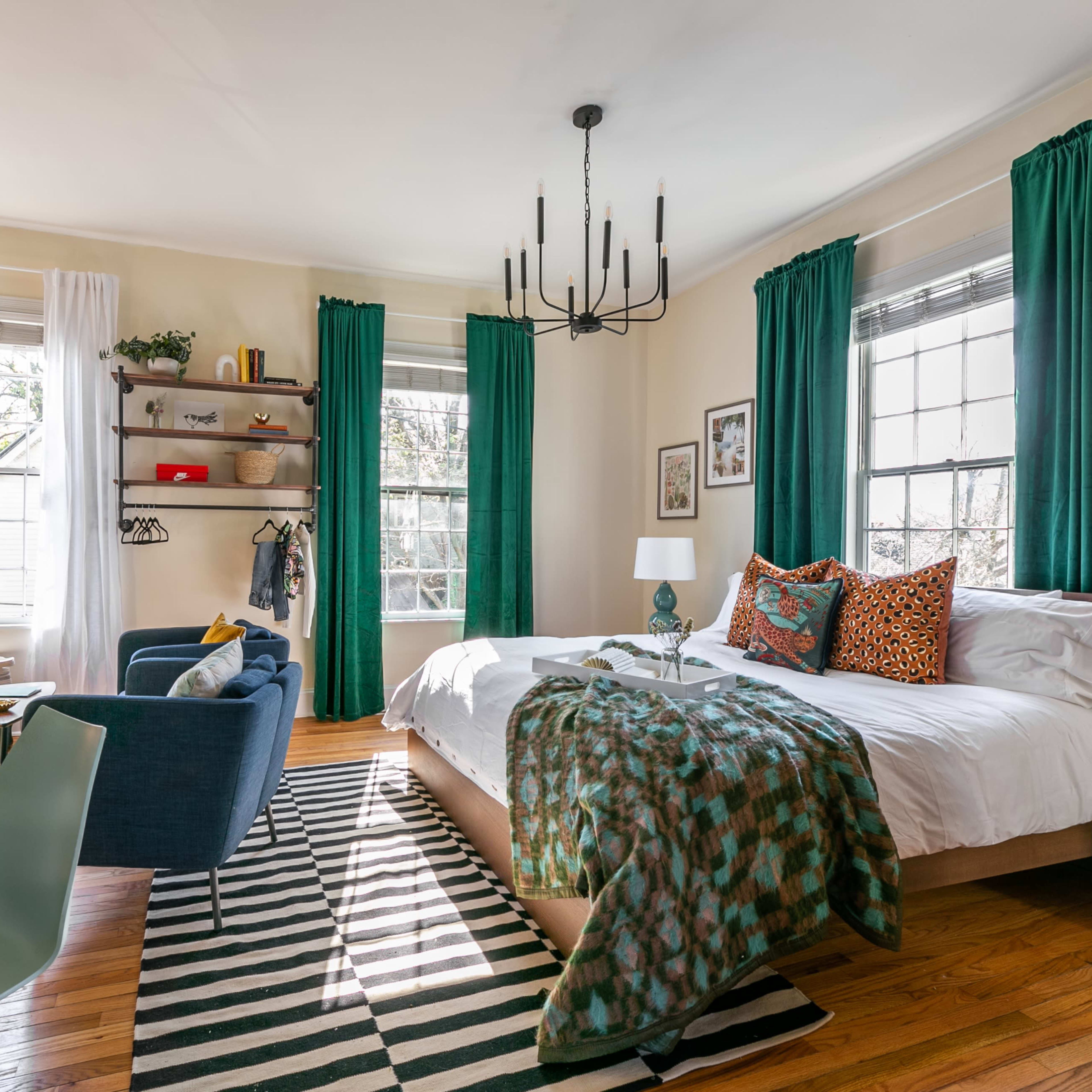 A bright bedroom featuring a bed with patterned pillows, a desk, a chair, and large windows draped with green curtains.