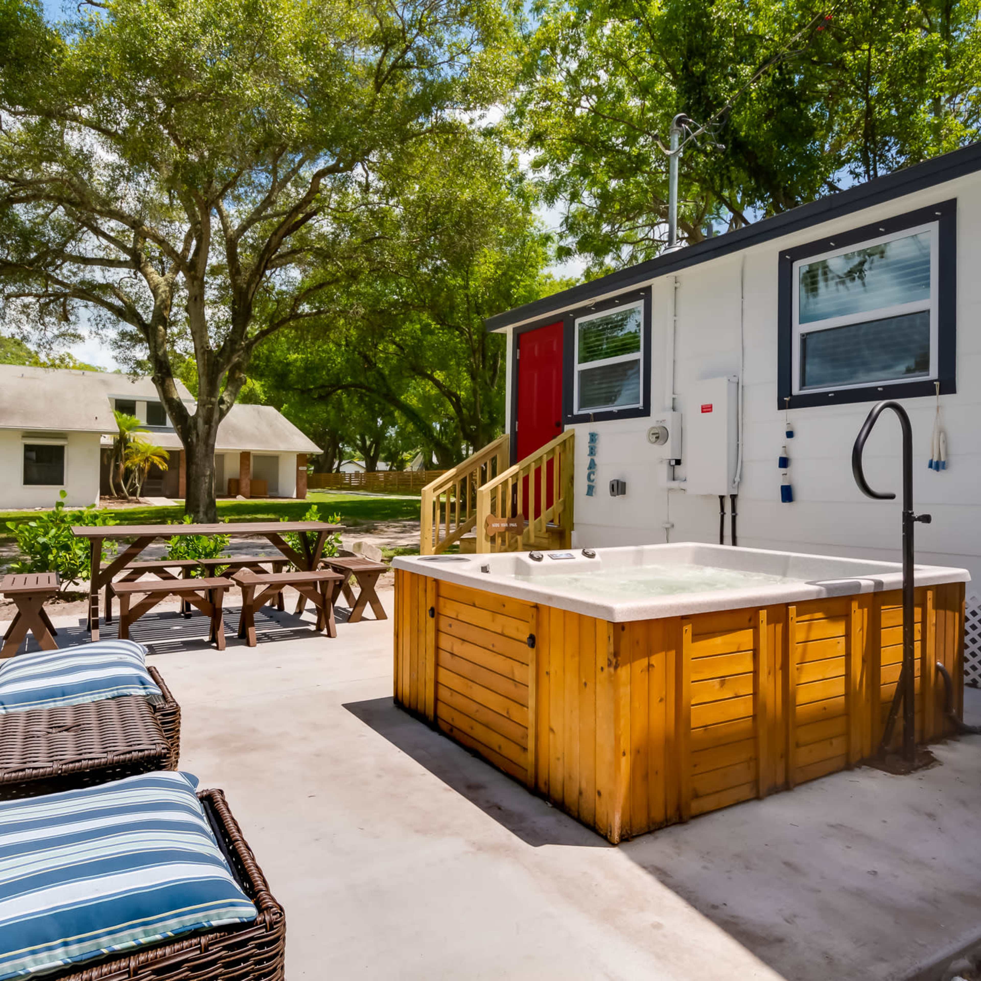 A hot tub is positioned on a patio next to a small seating area and a white building with a red door, surrounded by trees and another building in the background.