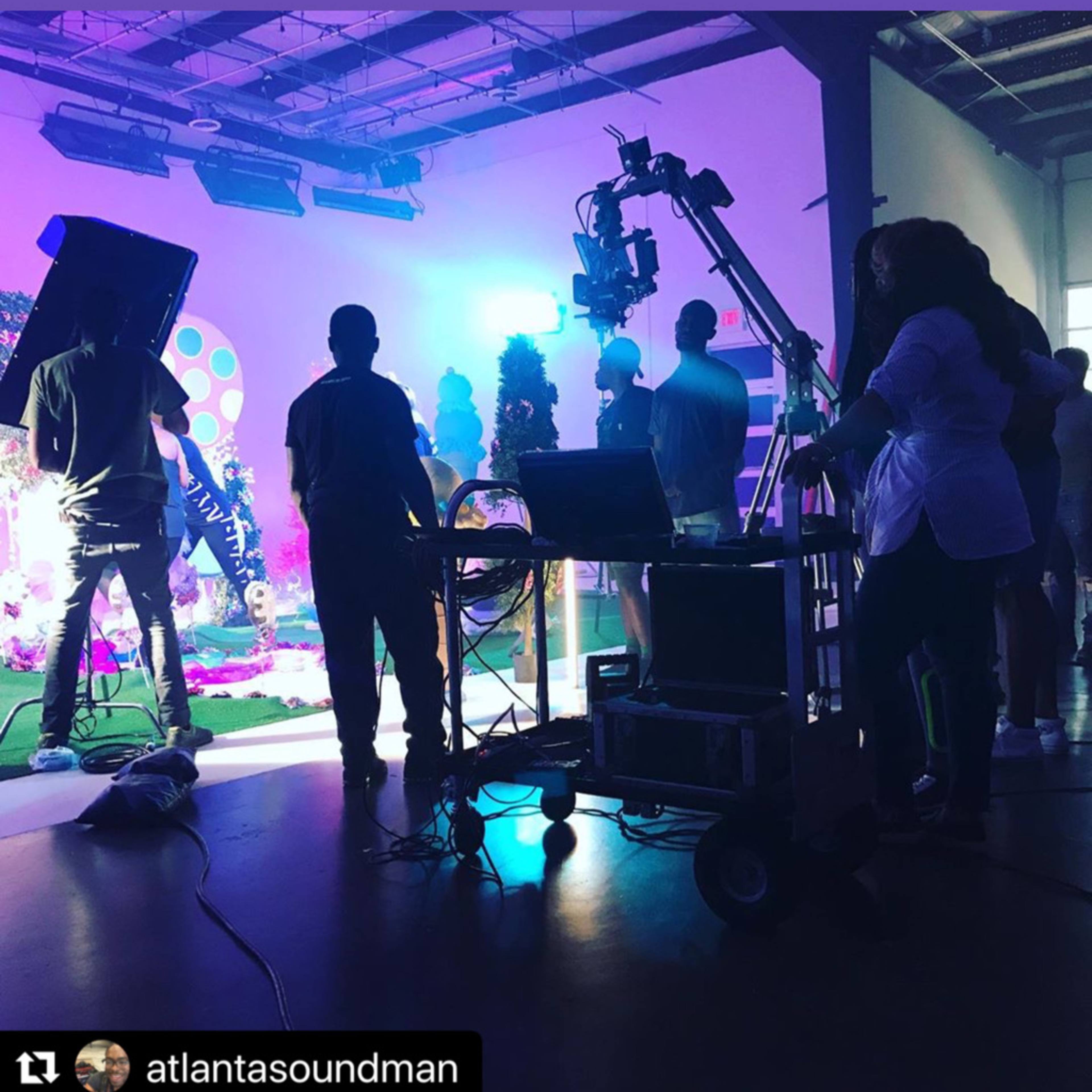 A production set with a crew preparing around a camera on a tripod, illuminated by colorful lights.
