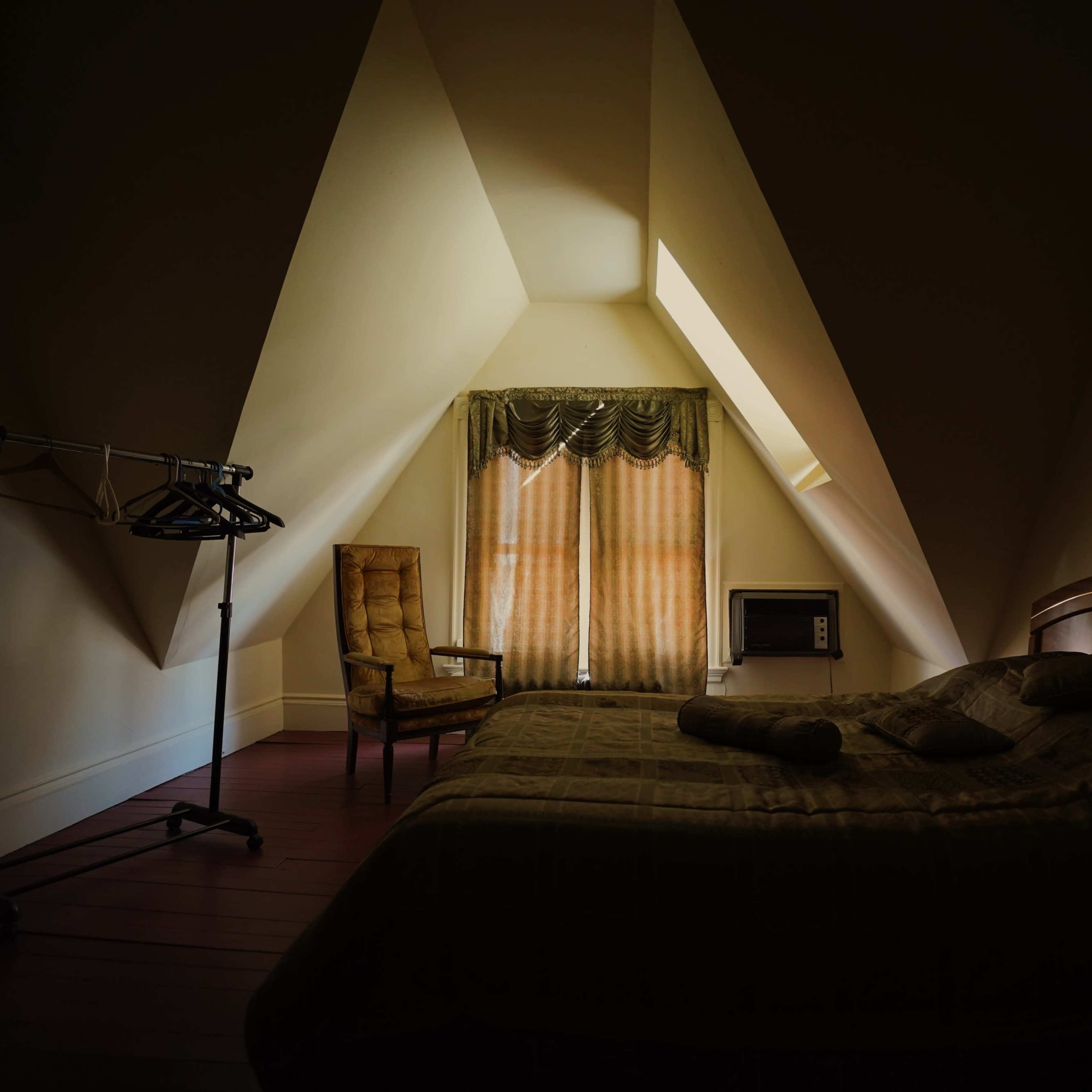 A sparsely furnished bedroom features a slanted ceiling, a bed with a patterned blanket, a chair, a clothing rack, and a window with draped curtains.