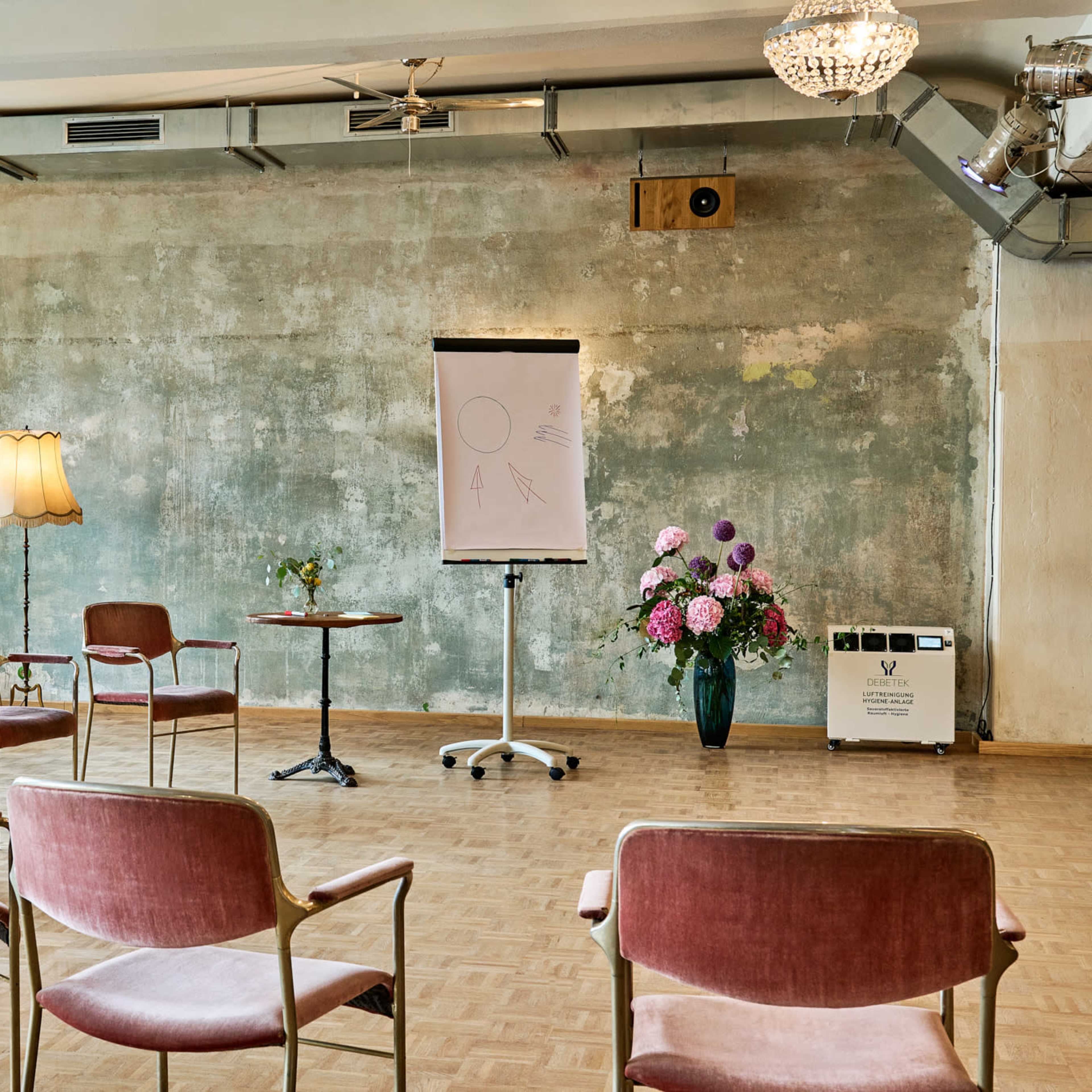 A spacious room features a set of chairs arranged around a central table, with a flip chart and potted plants against a textured wall.