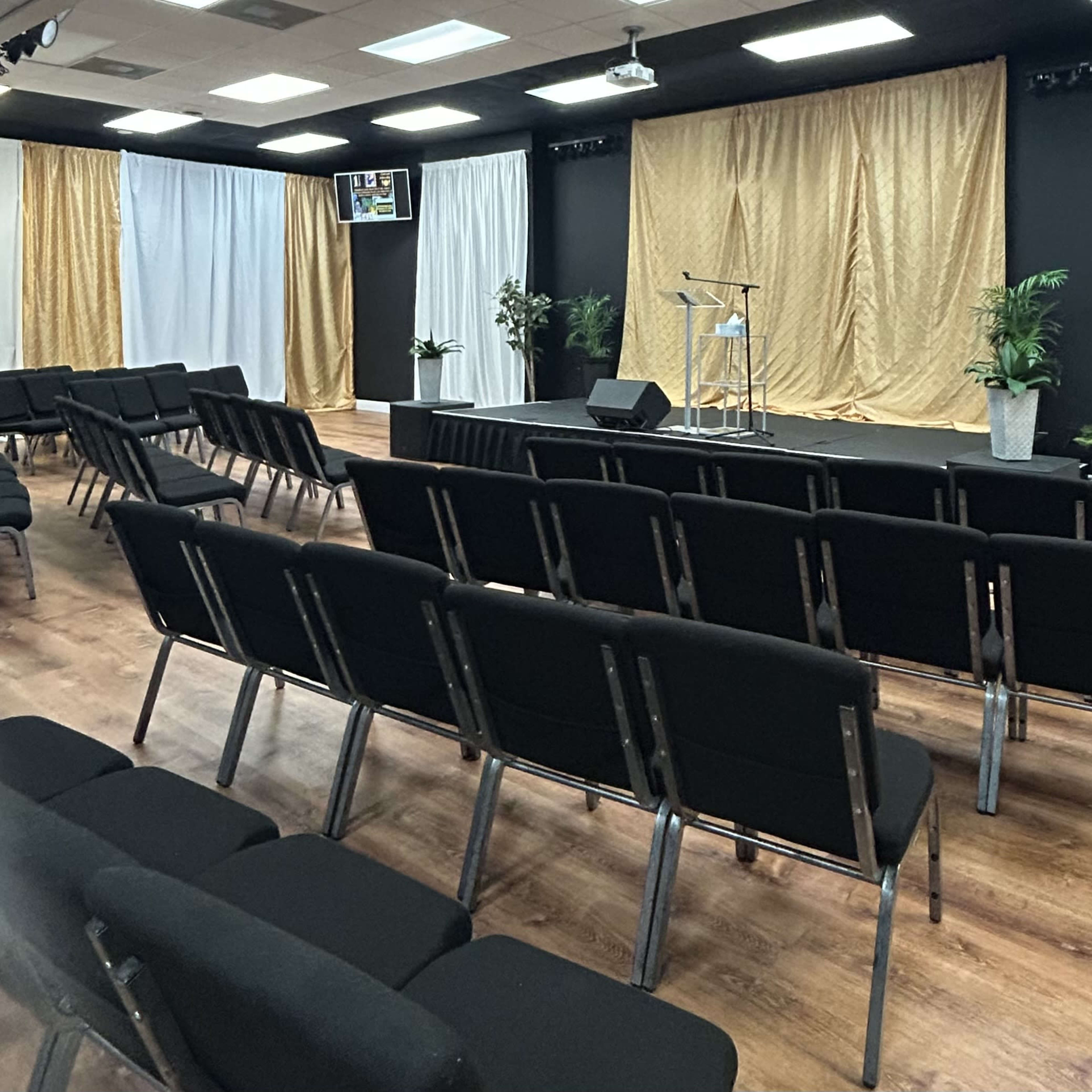 An auditorium-style room with black chairs arranged in rows facing a small stage decorated with white and gold drapes.