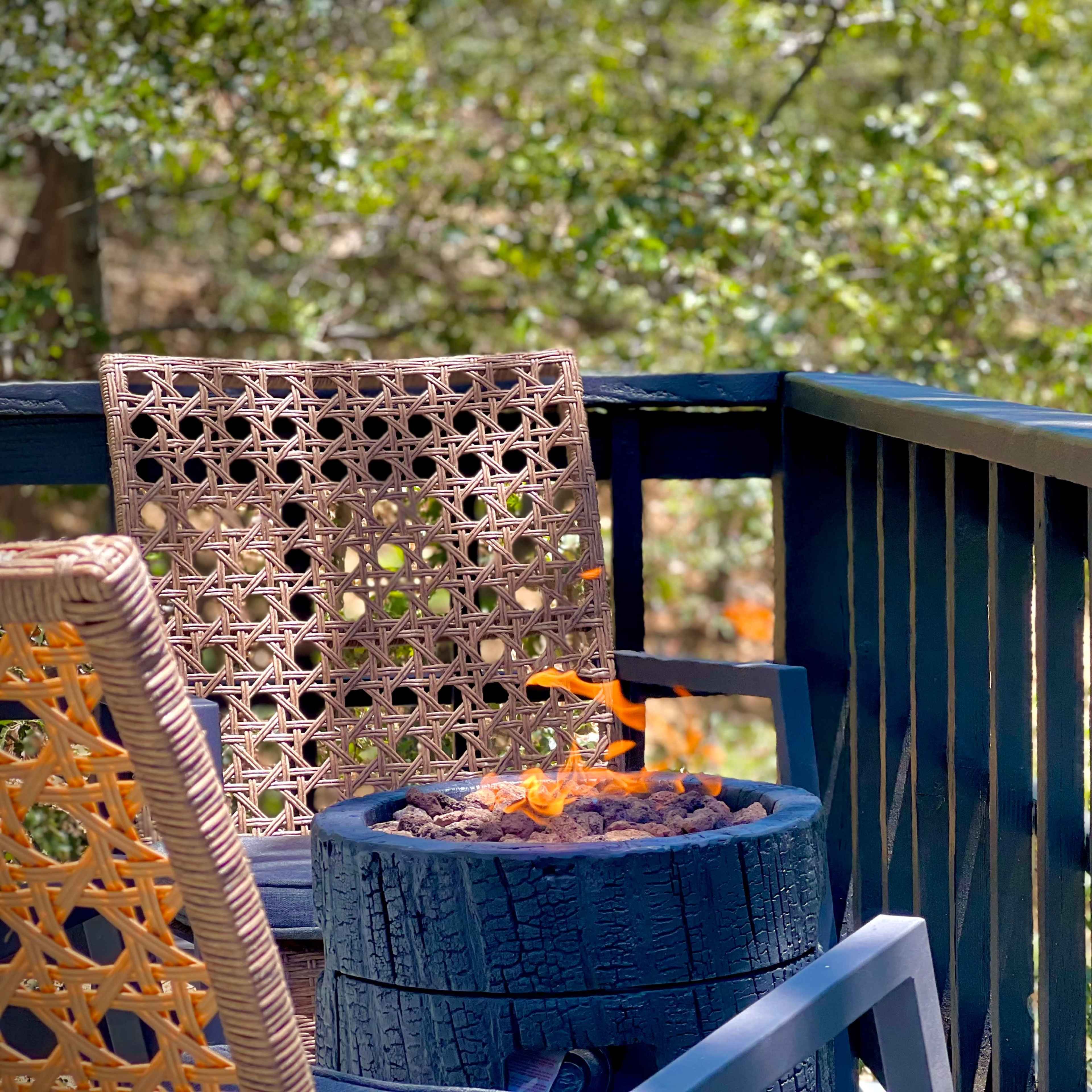 A wooden deck features a fire pit surrounded by a woven chair and two glasses of red wine placed on the railing.