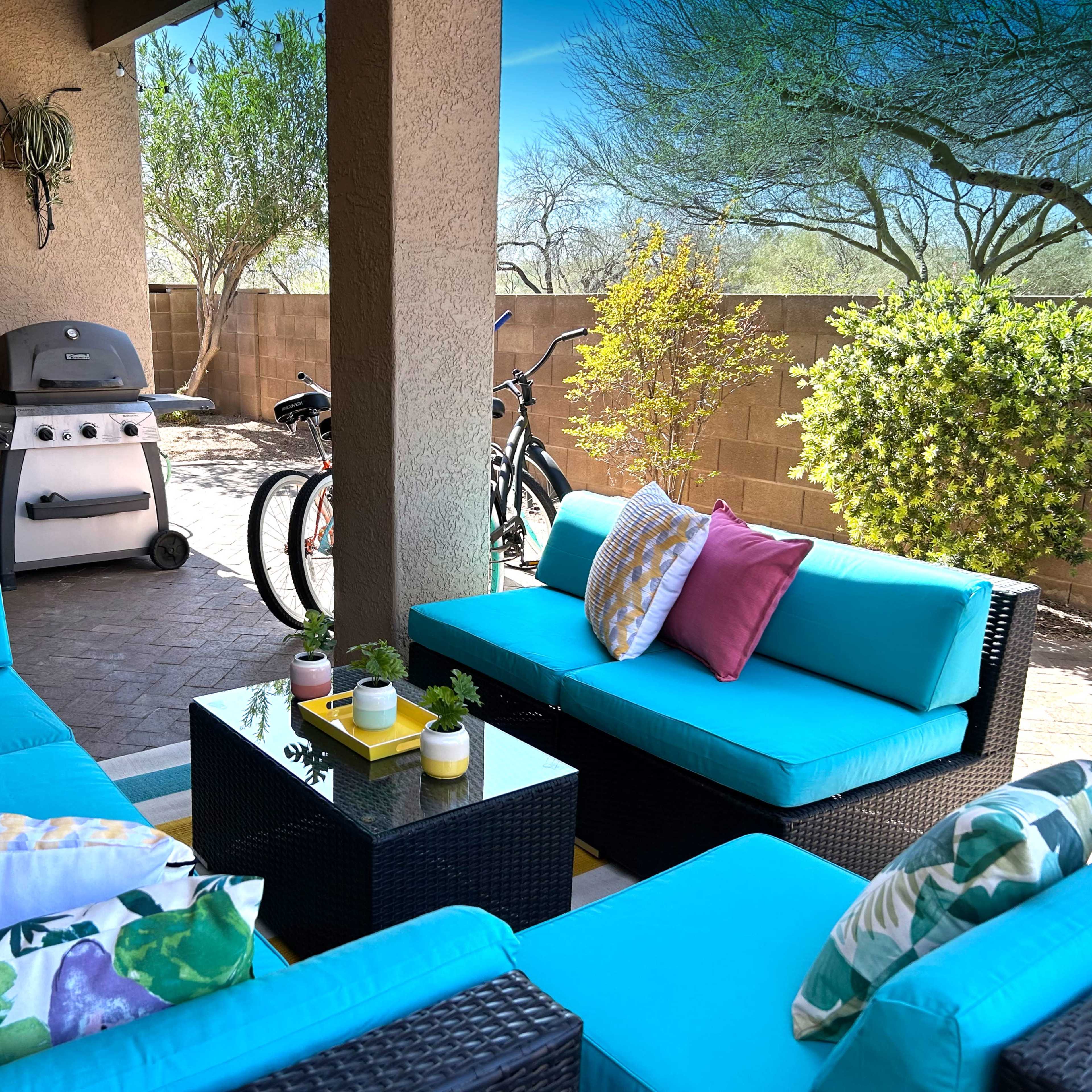 A patio area features turquoise outdoor furniture arranged around a small table, with a grill and bicycles nearby against a backdrop of plants and a wall.