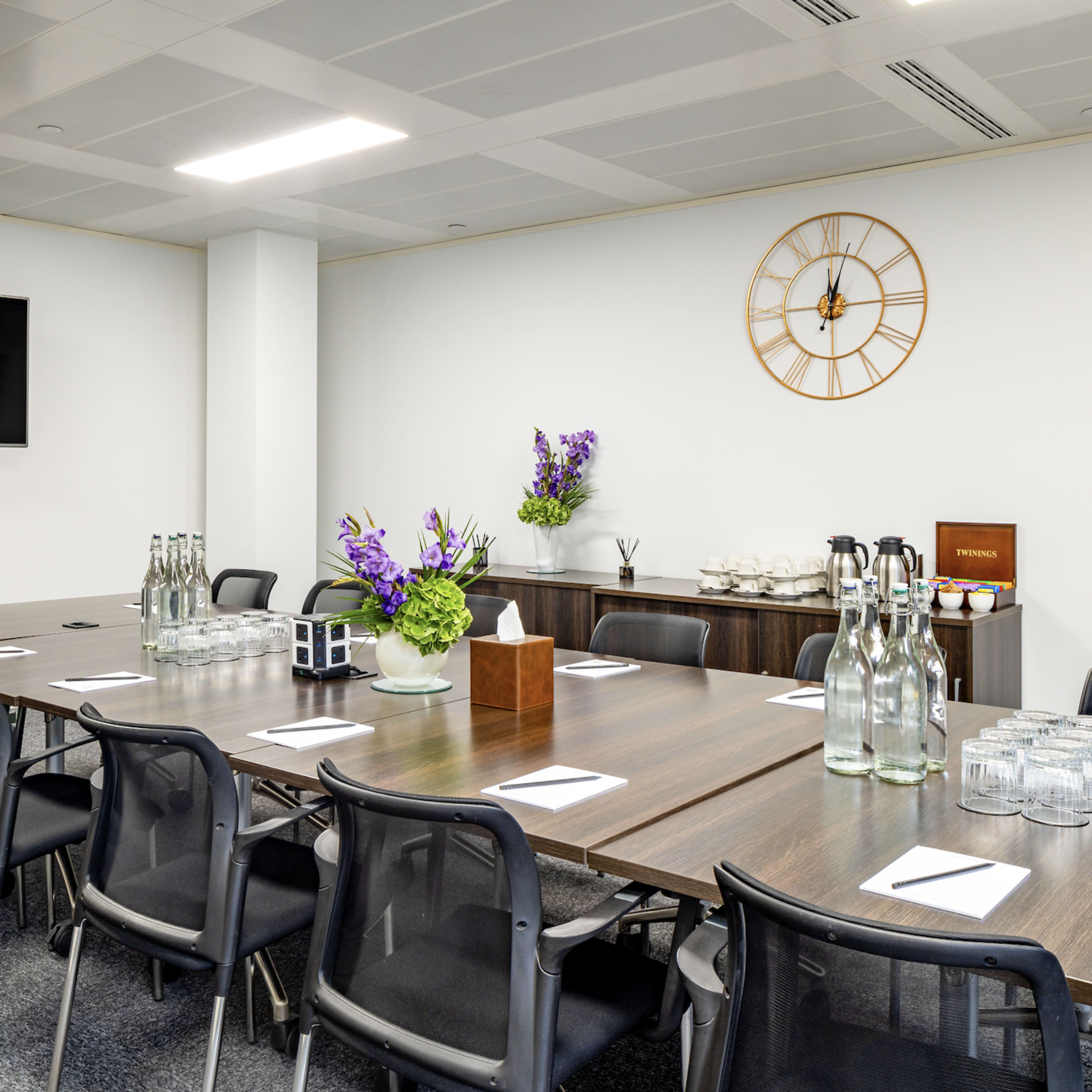 A modern conference room features a long table surrounded by chairs, with a television mounted on the wall and a clock above a sideboard.