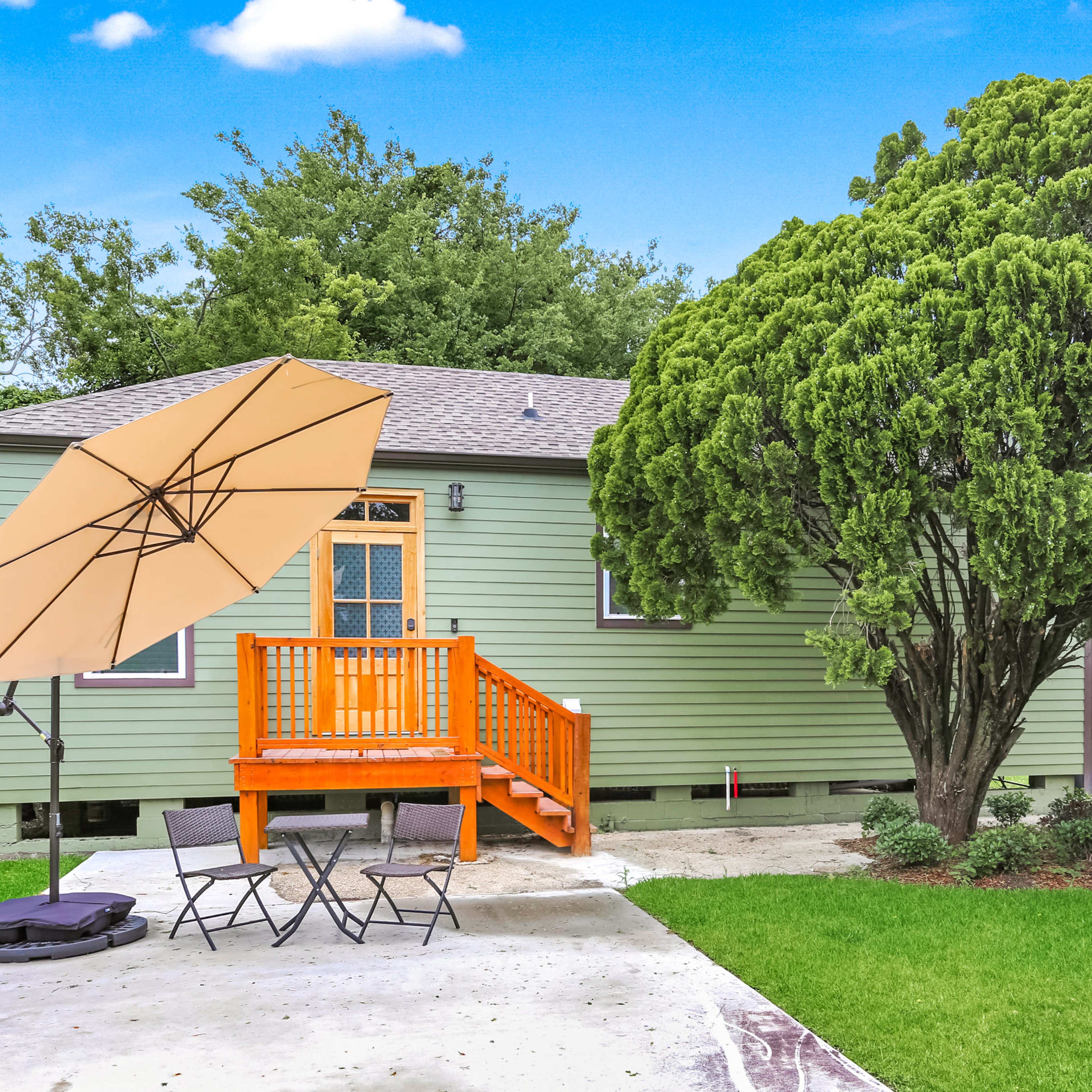 A green house with a wooden deck and orange umbrella, surrounded by a well-maintained yard with a large tree.
