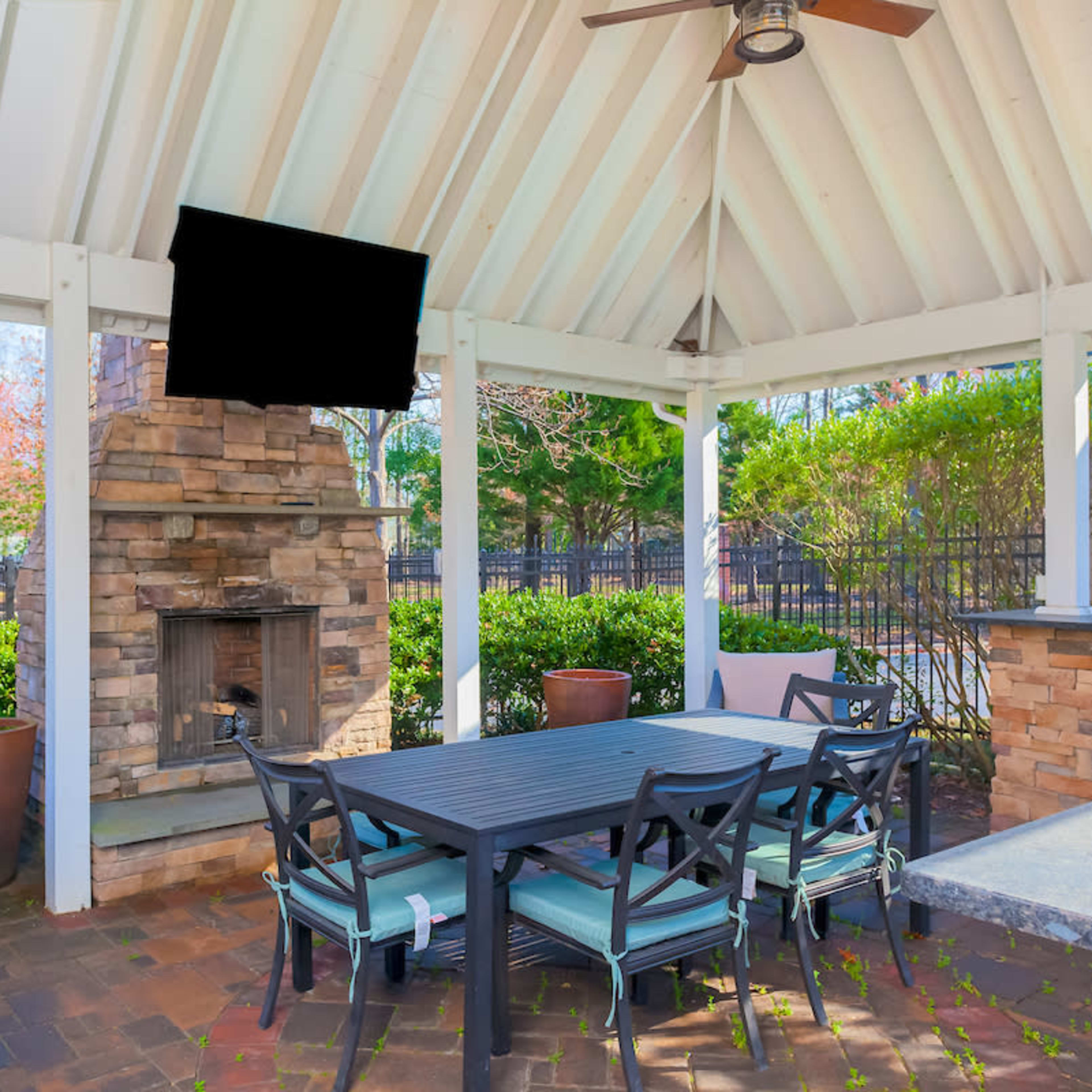 A covered outdoor patio features a stone fireplace and a dining table with chairs, surrounded by greenery and a barbecue grill.