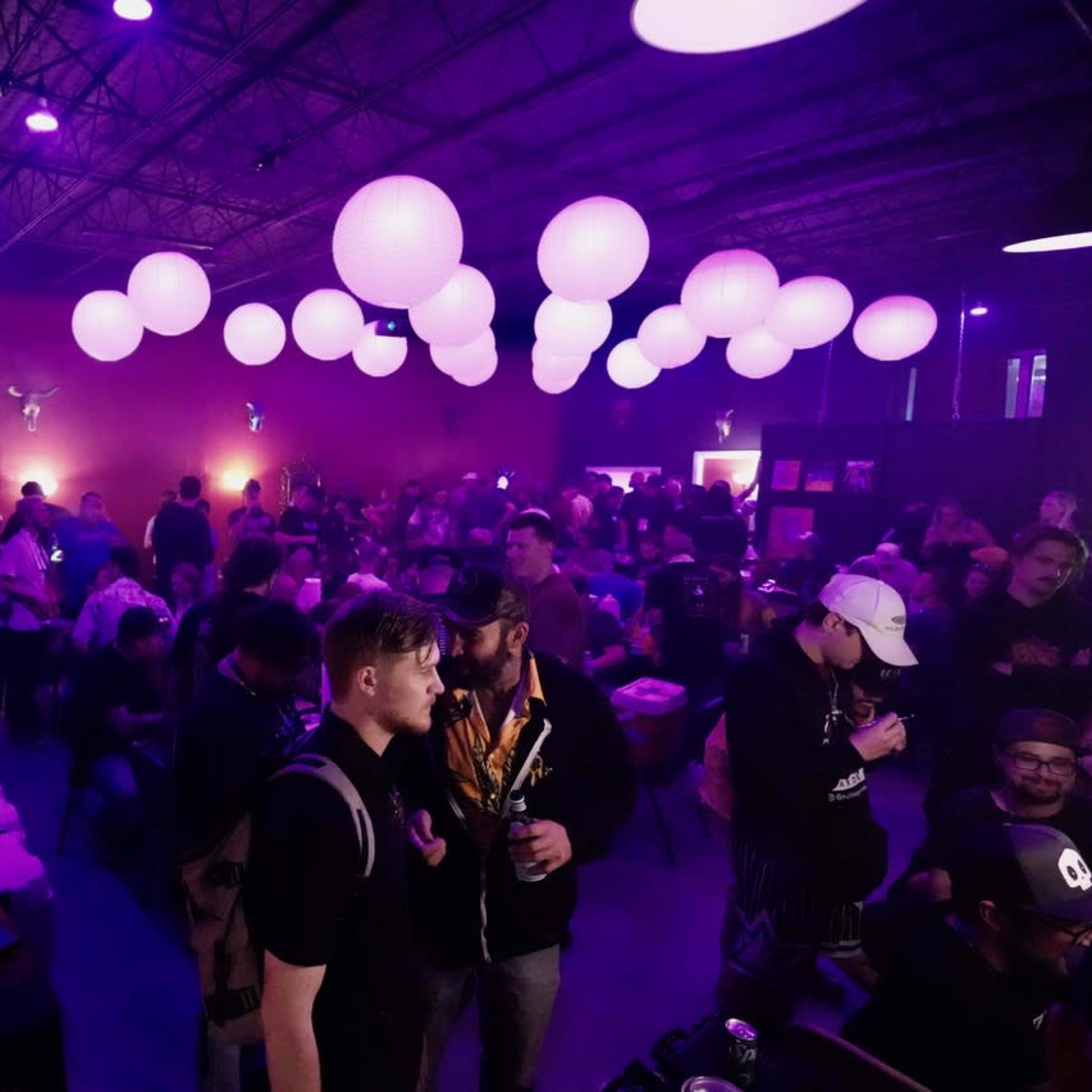 A crowded event space features people socializing under large, glowing white lanterns and a DJ setup in the corner.