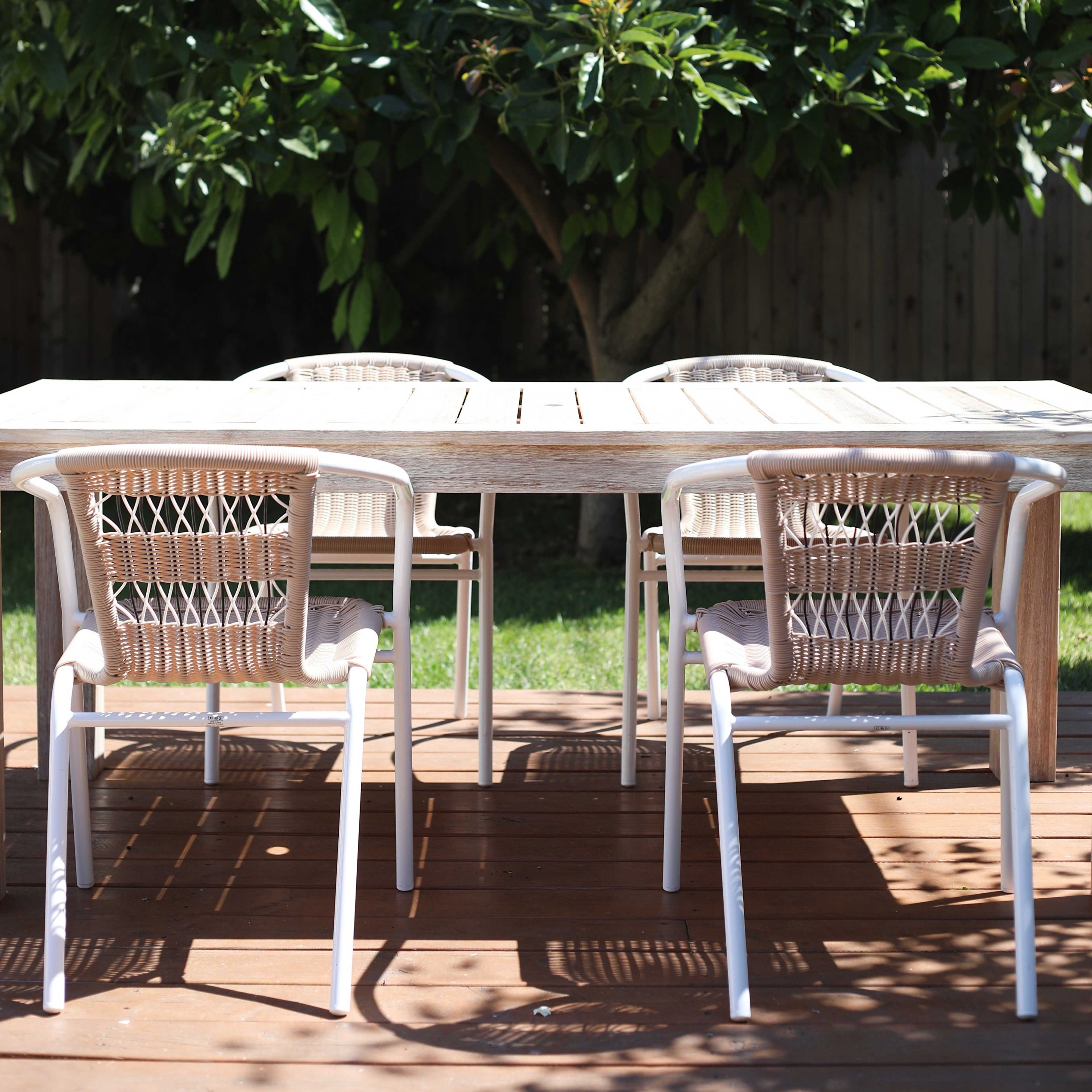 A wooden dining table is surrounded by four chairs on a deck beneath a tree.