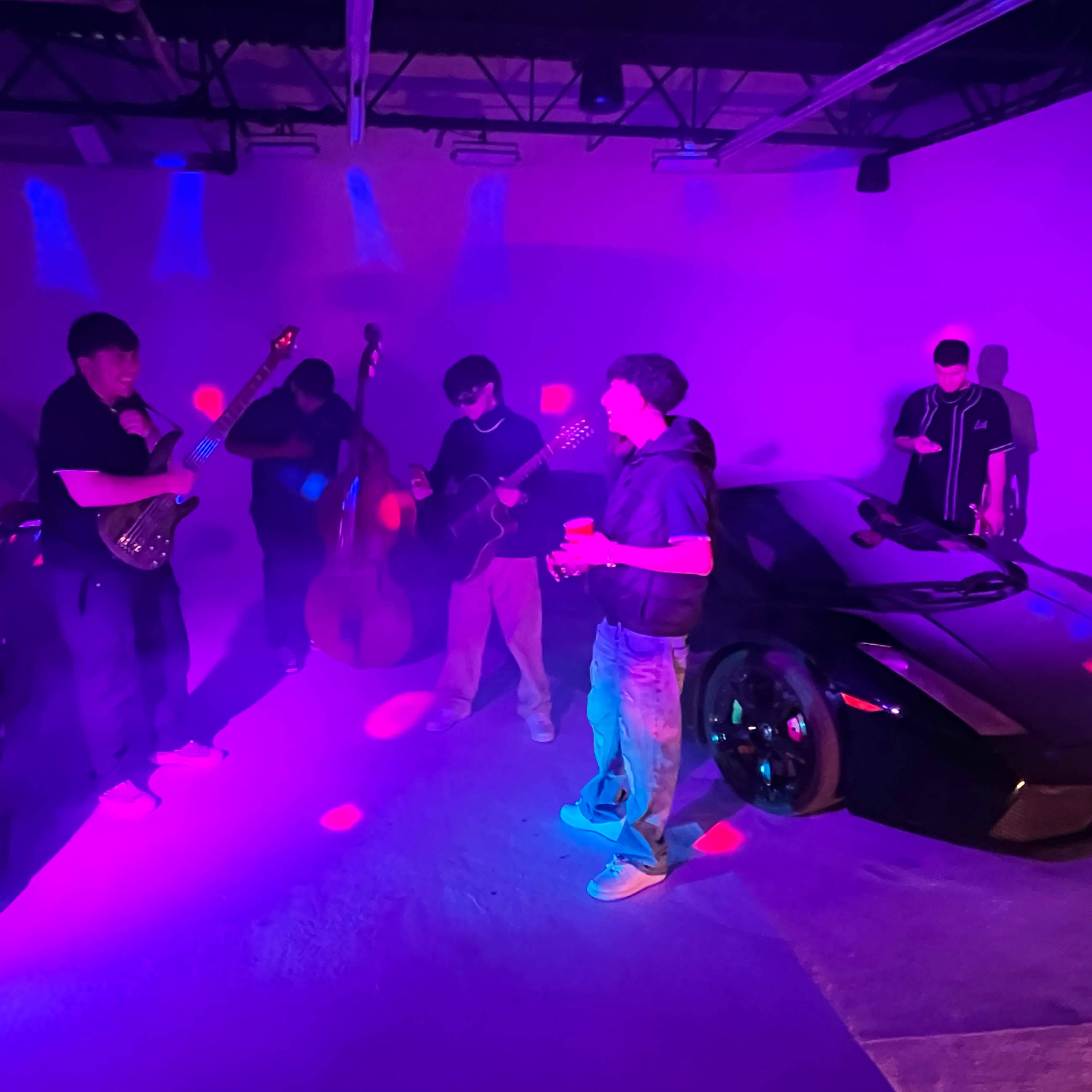 A group of musicians is playing instruments near two luxury cars in a dimly lit space with purple and blue lighting.