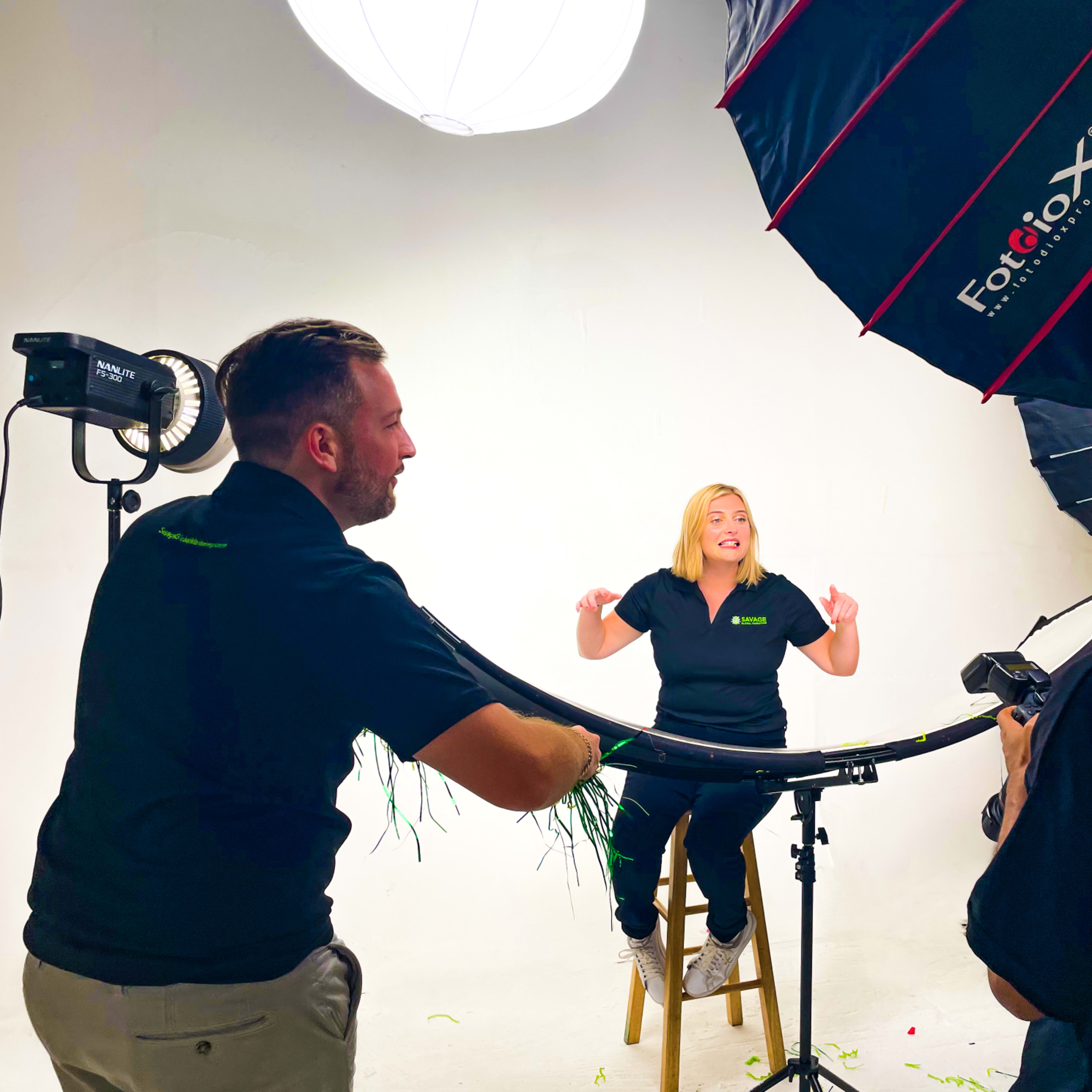 A group of photographers is setting up a studio shoot with a woman sitting on a stool against a white backdrop.