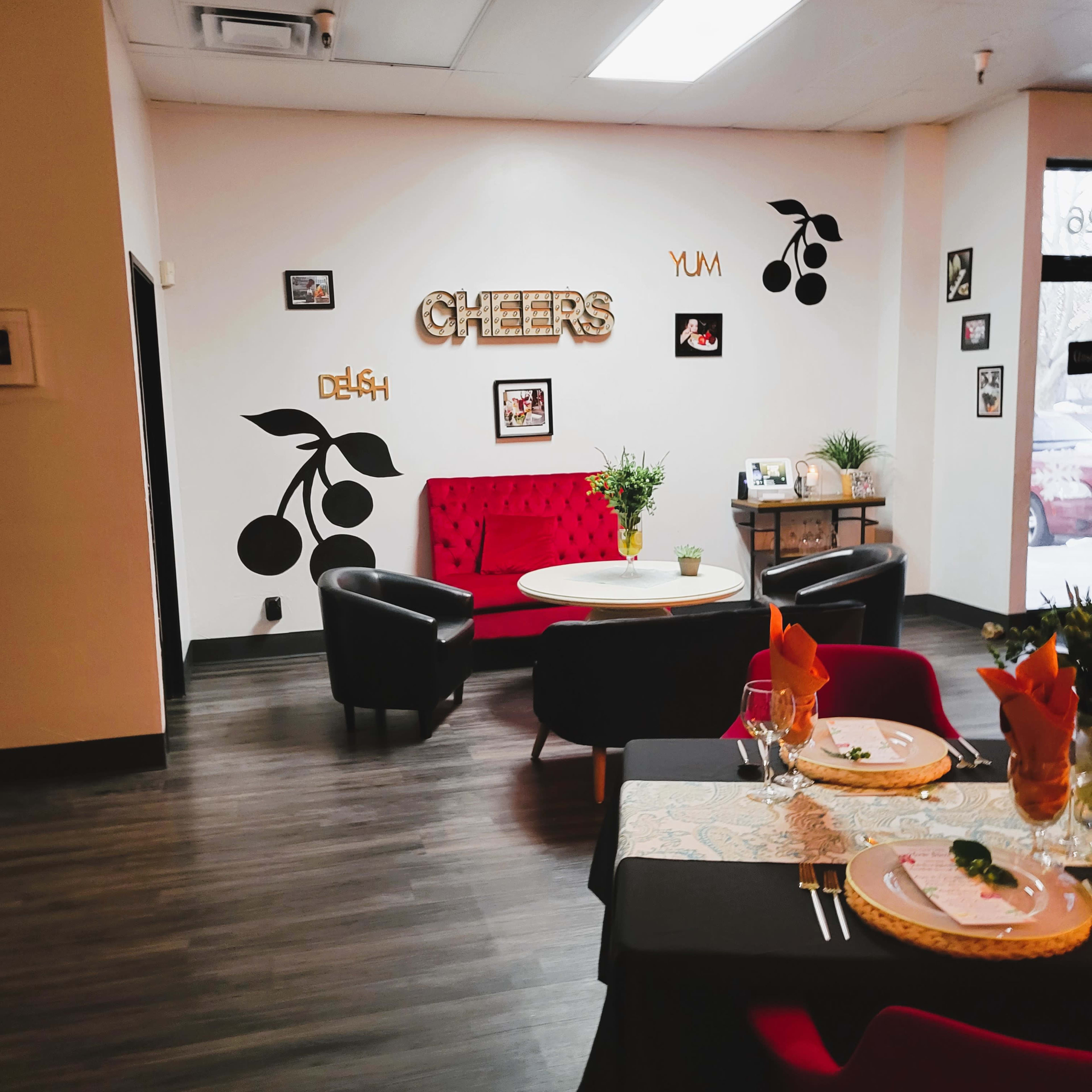 A cozy dining area features a red velvet sofa, elegant tables set with flowers, and wall decorations that include the words "CHEERS" and cherry motifs.