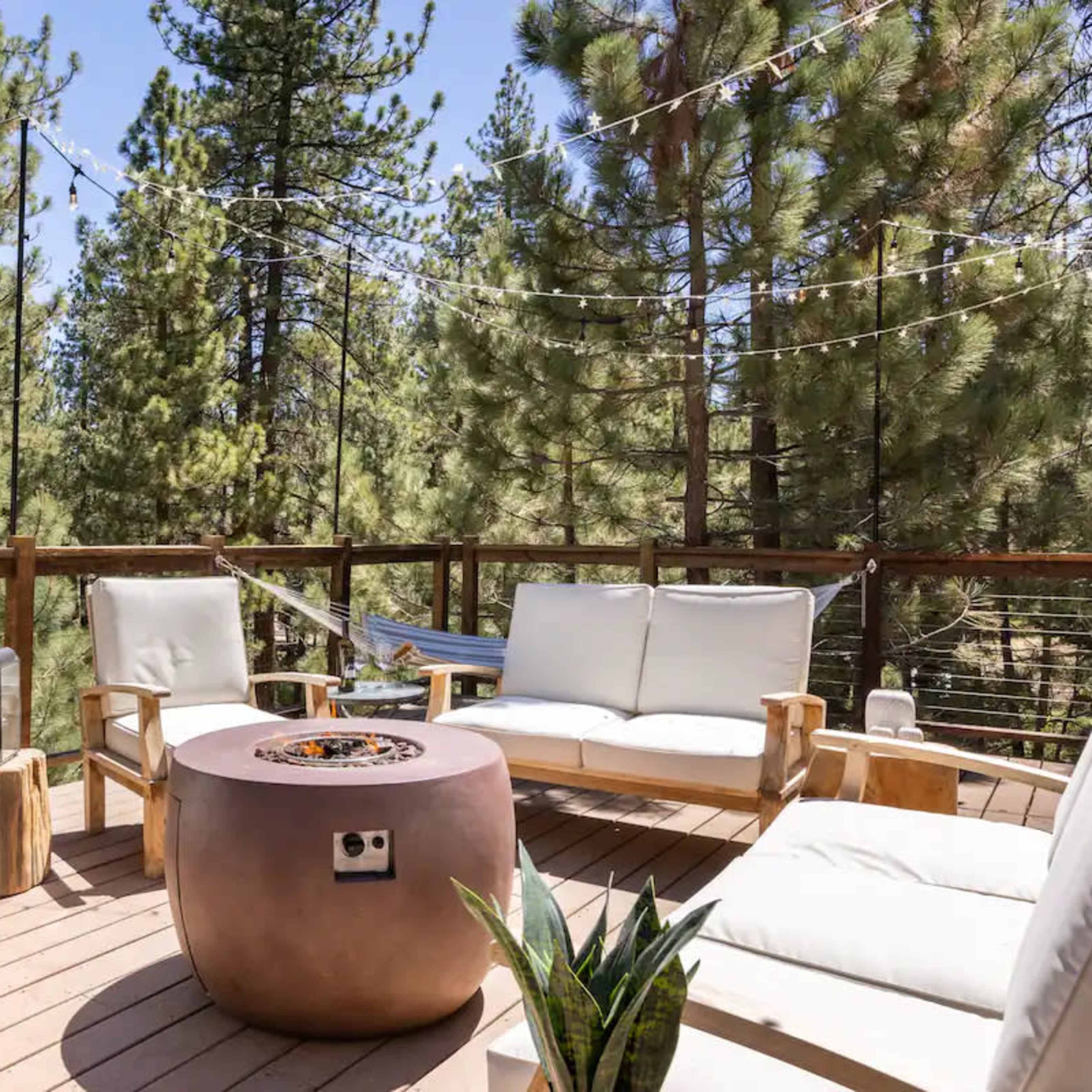 A wooden deck features a circular fire pit surrounded by white chairs, with tall pine trees in the background.