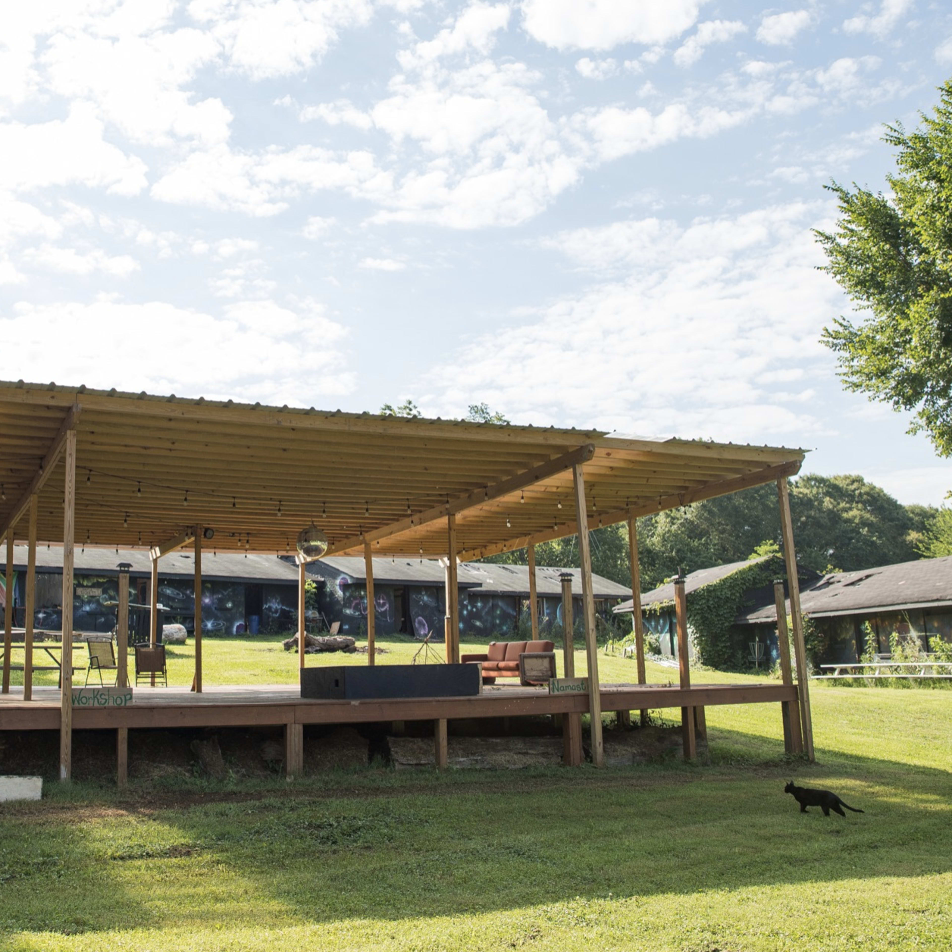 A wooden pavilion with a slatted roof stands on a grassy area, with benches and a small stage in front, and a black cat walking nearby.