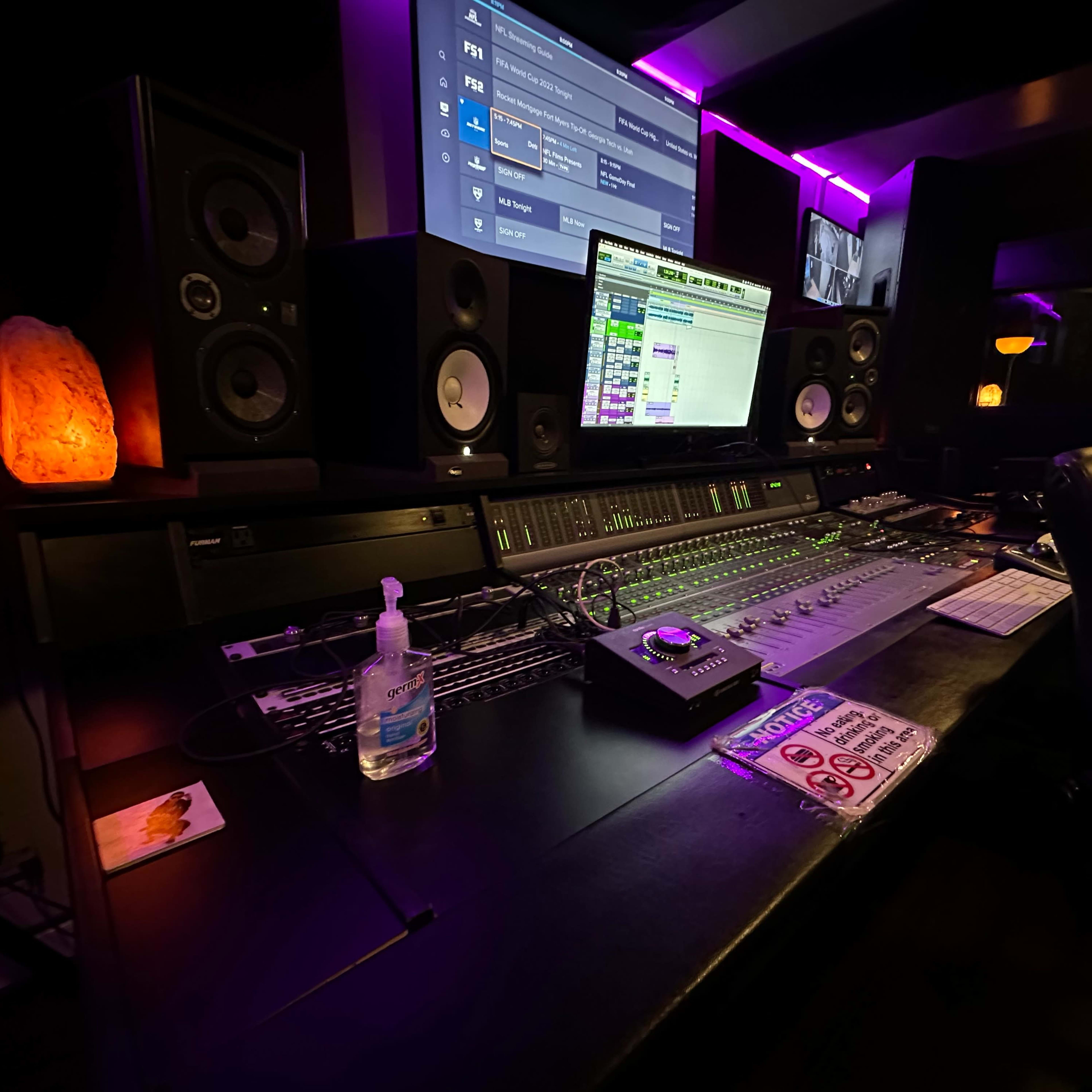 A recording studio control desk with a mixing console, computer monitor, and sound equipment, illuminated by purple lighting.