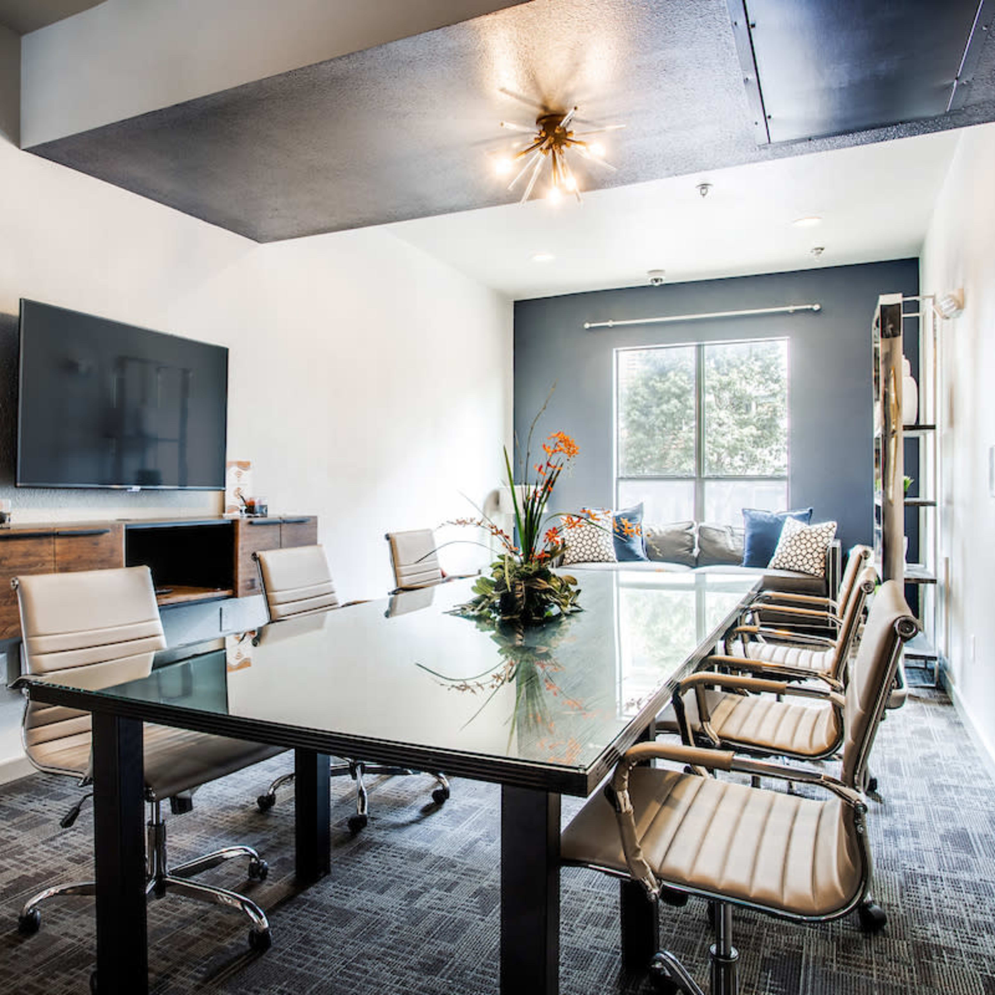 A modern conference room features a glass table surrounded by eight chrome chairs, with a large television mounted on the wall and a seating area in the background.