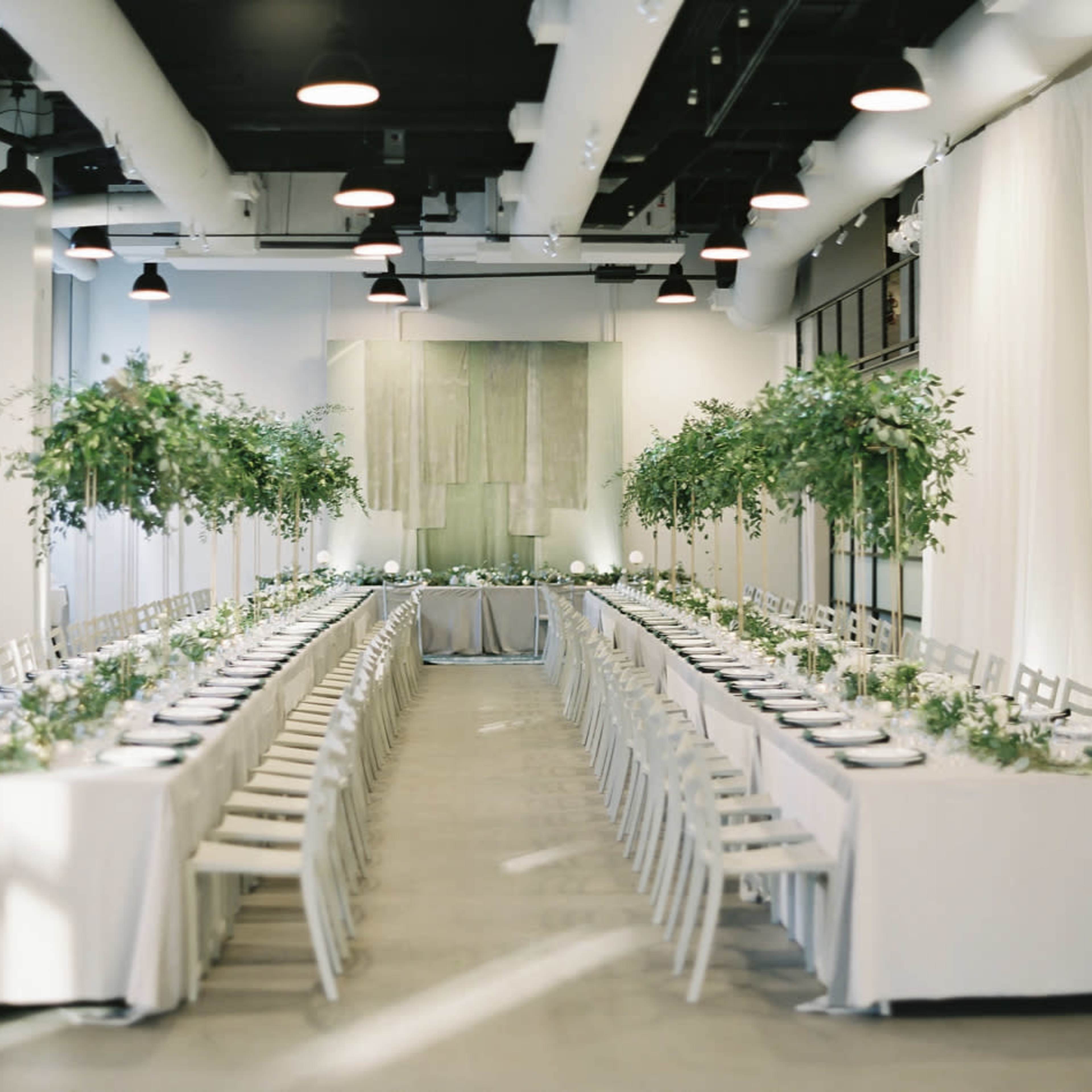 A long, elegantly set dining table is arranged in a spacious hall, flanked by tall greenery and white tablecloths.