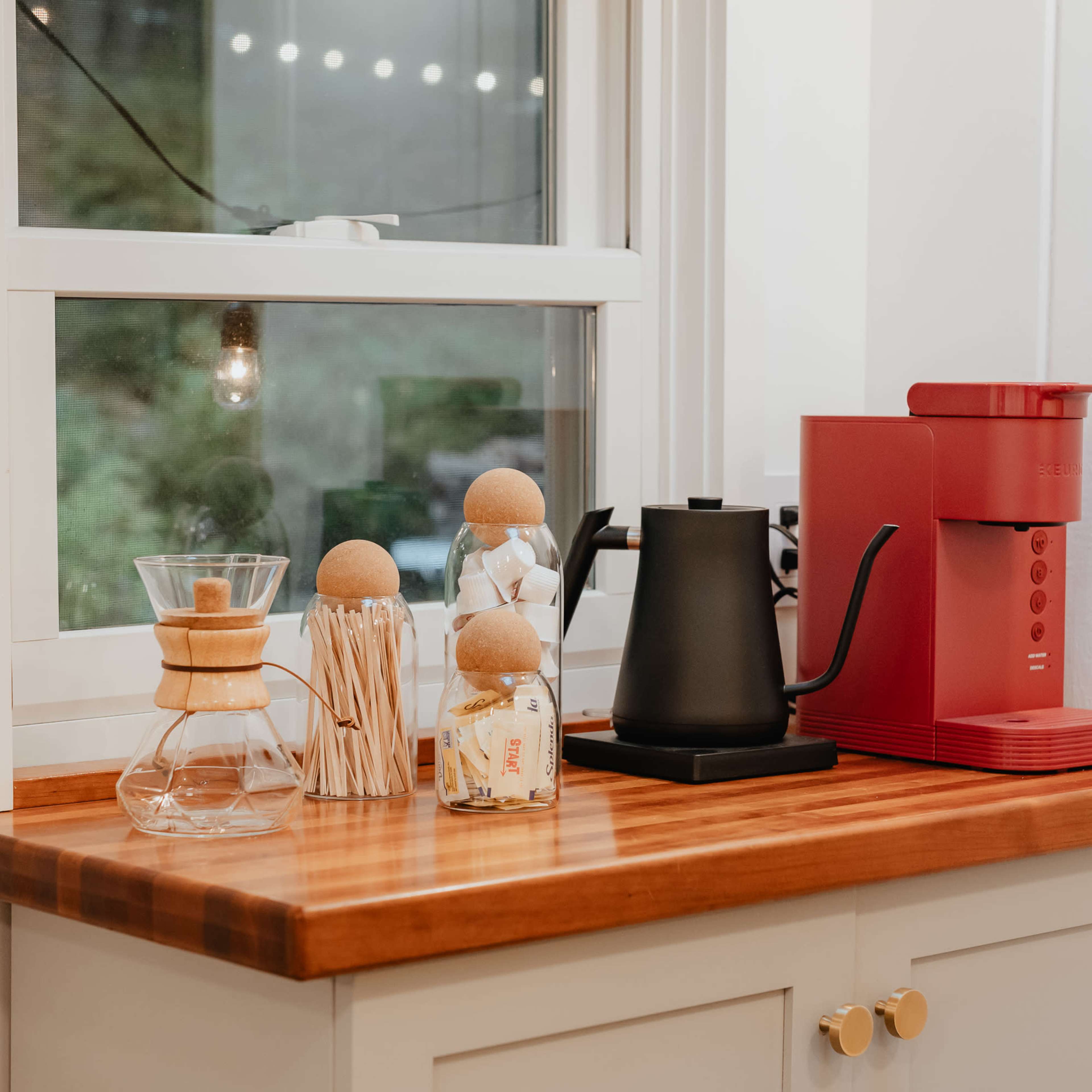 A wooden countertop holds a red coffee machine, a black kettle, a glass coffee maker, and decorative items near a window.