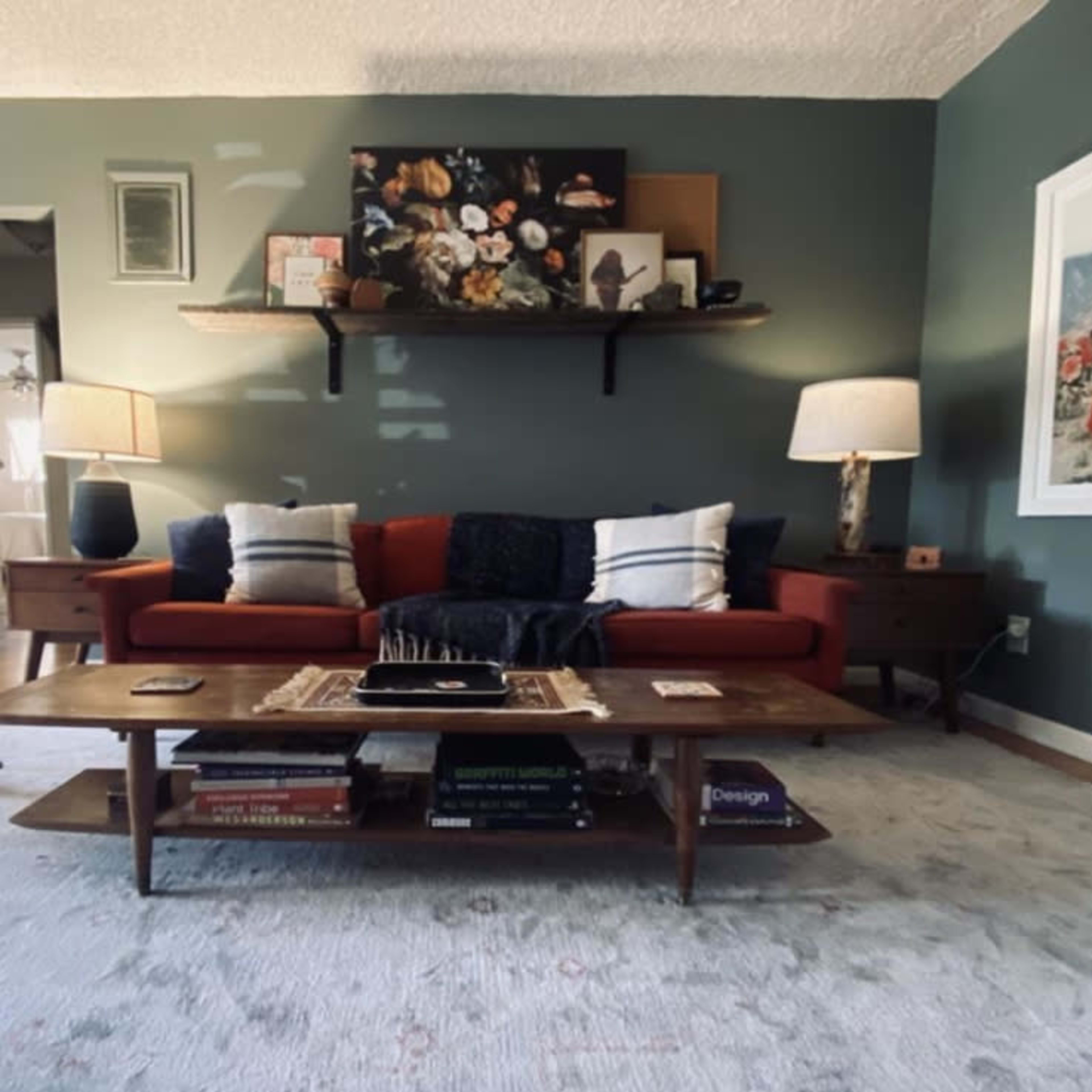The living room features a red sofa with blue and white throw pillows, a wooden coffee table stacked with books, and two lamps beside the sofa, all against a green wall.