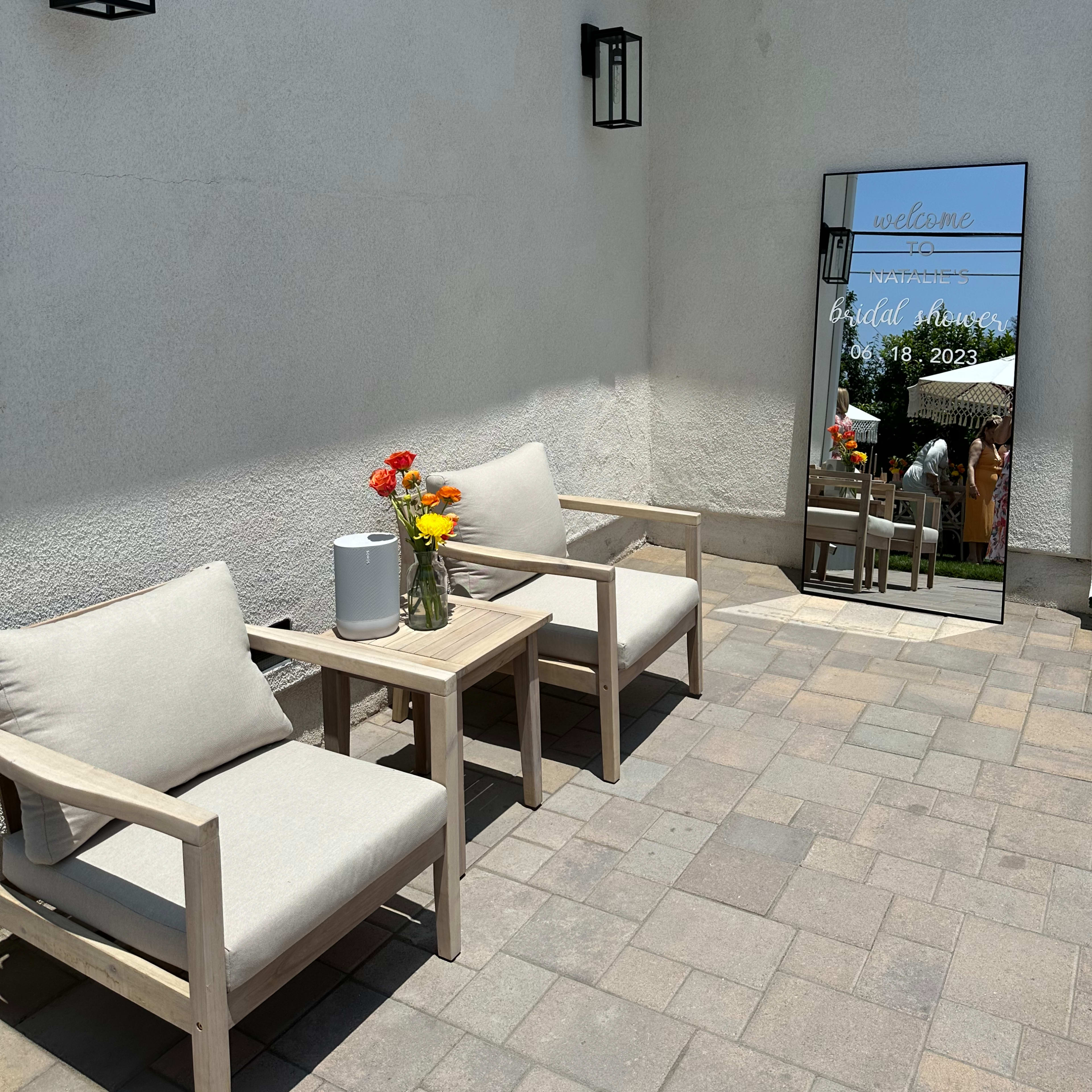 A sunlit outdoor space featuring two wooden chairs with cushions, a small table with a vase of flowers, and a large mirror reflecting a welcoming message and a shaded area in the background.