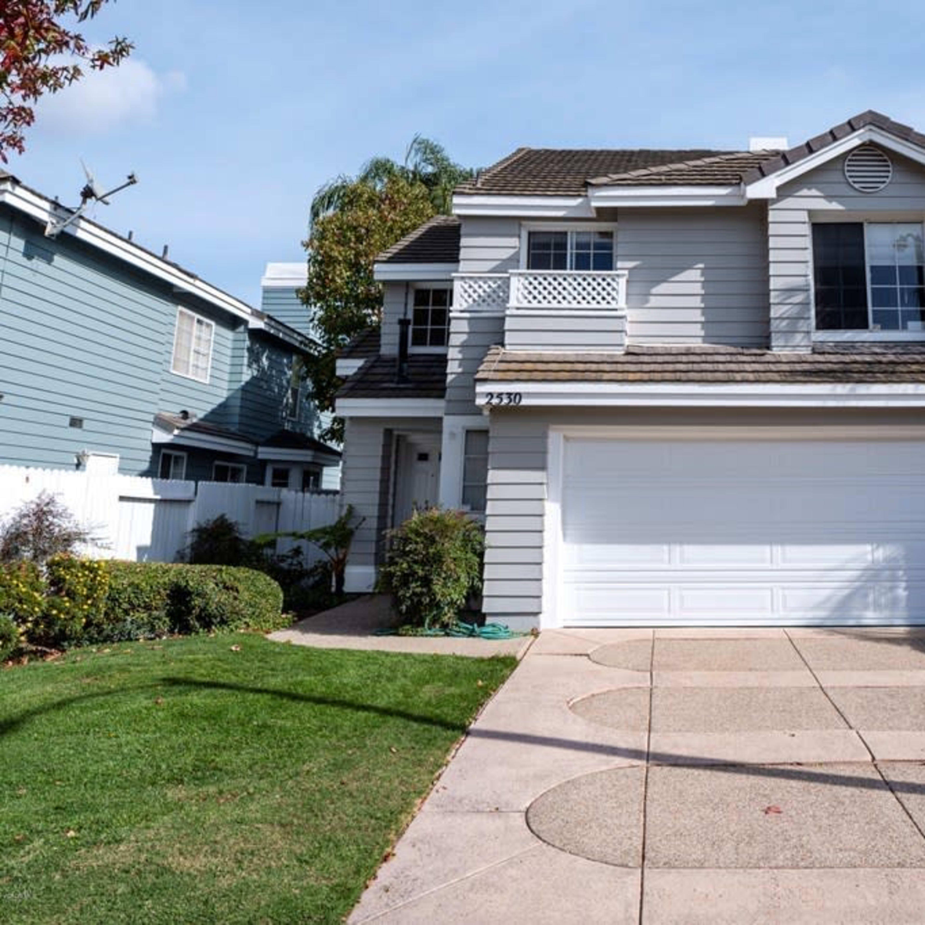 A two-story house with a gray exterior, a white garage door, and palm trees in the front yard.