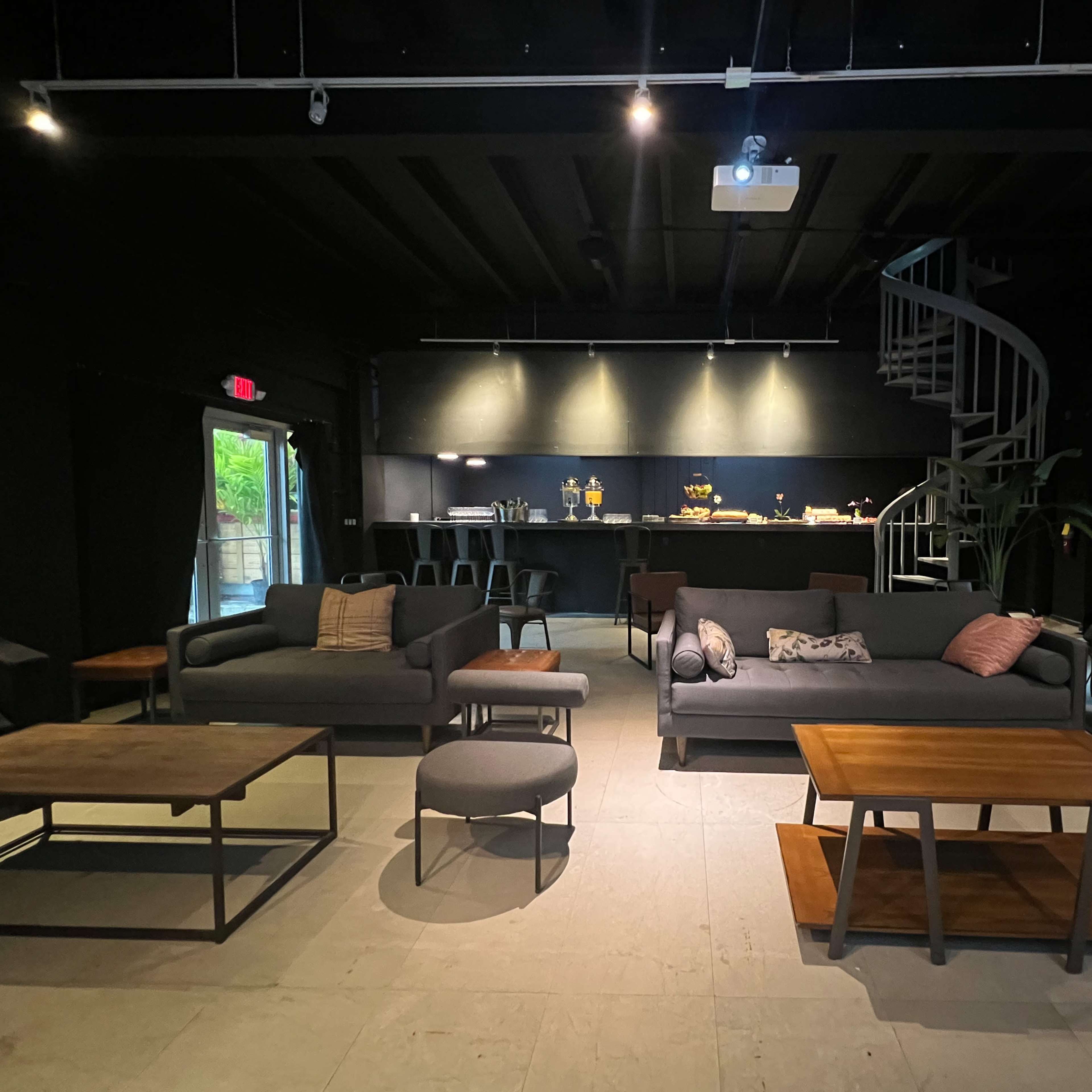 A modern lounge area with dark walls, featuring multiple seating arrangements, a spiral staircase, and a kitchen bar in the background.