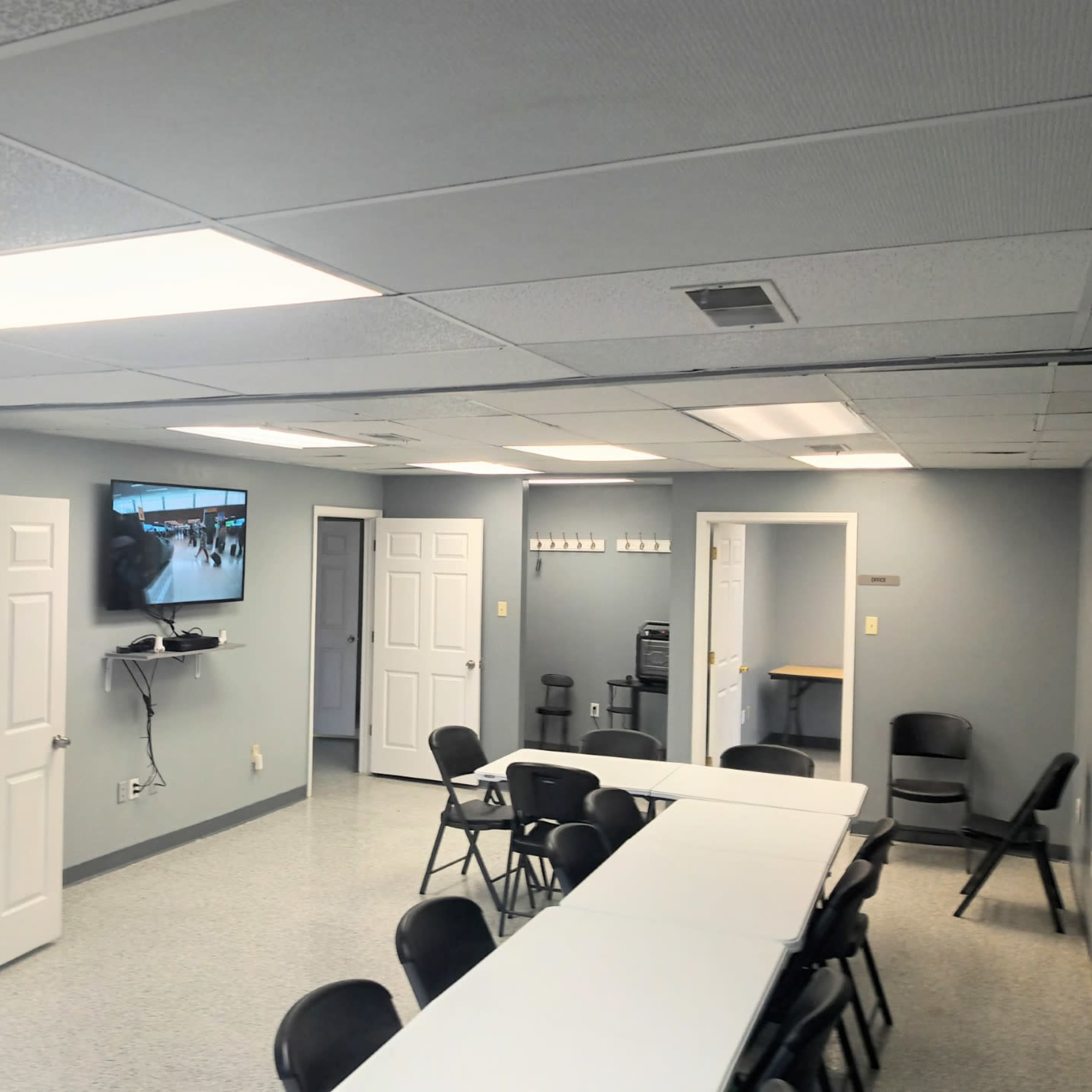 A spacious room with a long white table and black chairs, a TV mounted on the wall, and several doors leading to other areas.