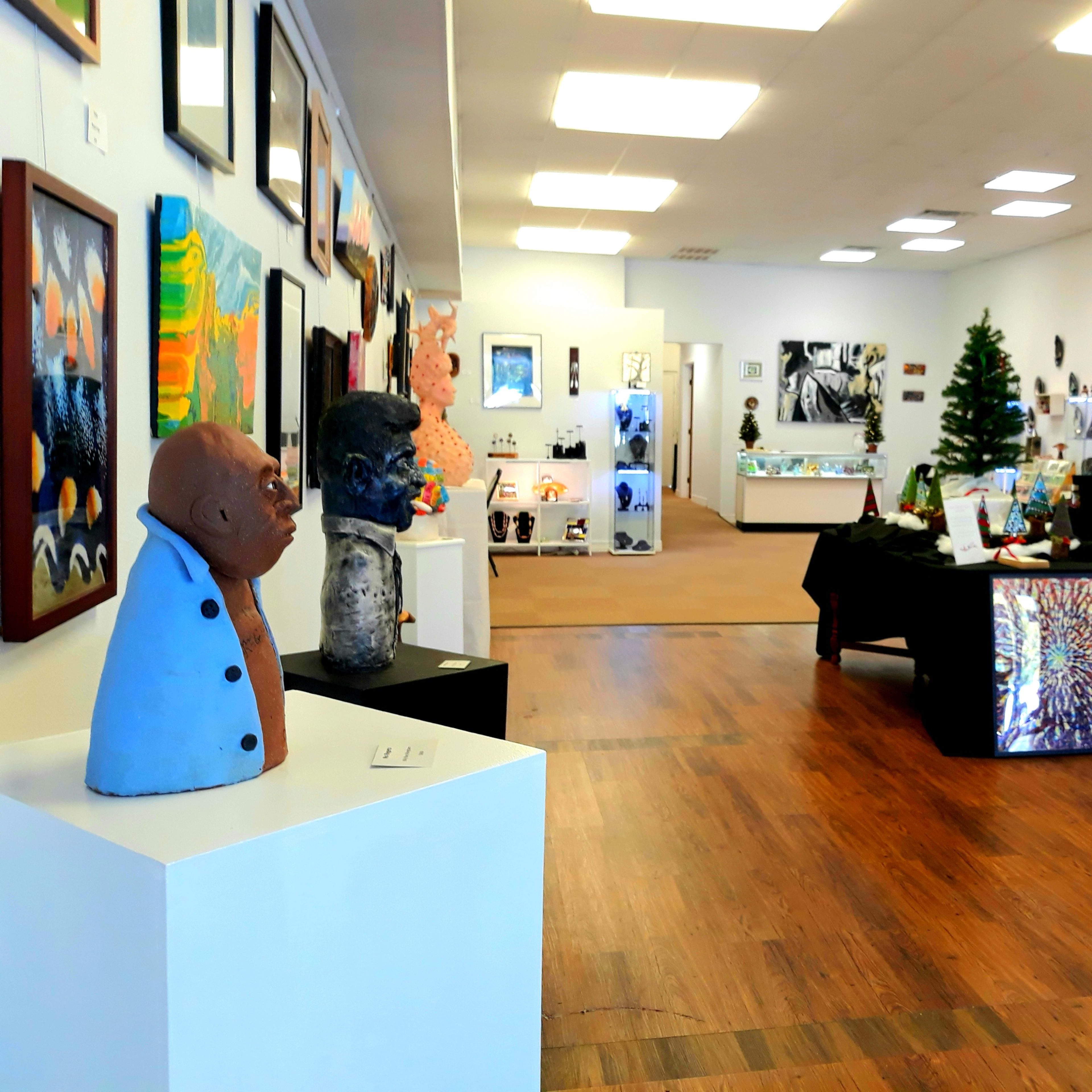 An art gallery featuring various sculptures and paintings displayed on white walls and wooden floors, with a Christmas tree and tables for additional artwork.