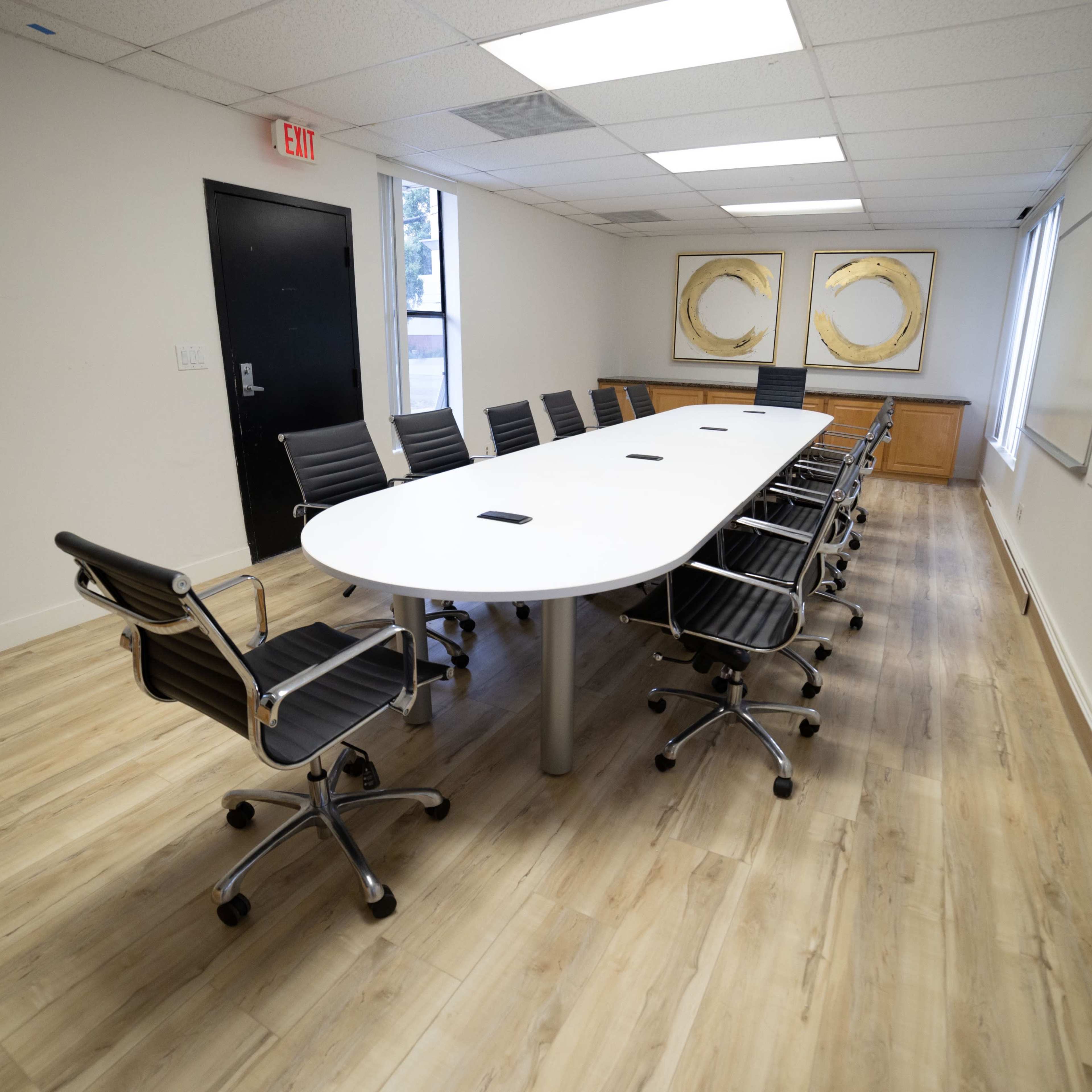 A conference room with a long white table surrounded by ten black ergonomic chairs, a whiteboard on one wall, and two circular artworks on another wall.