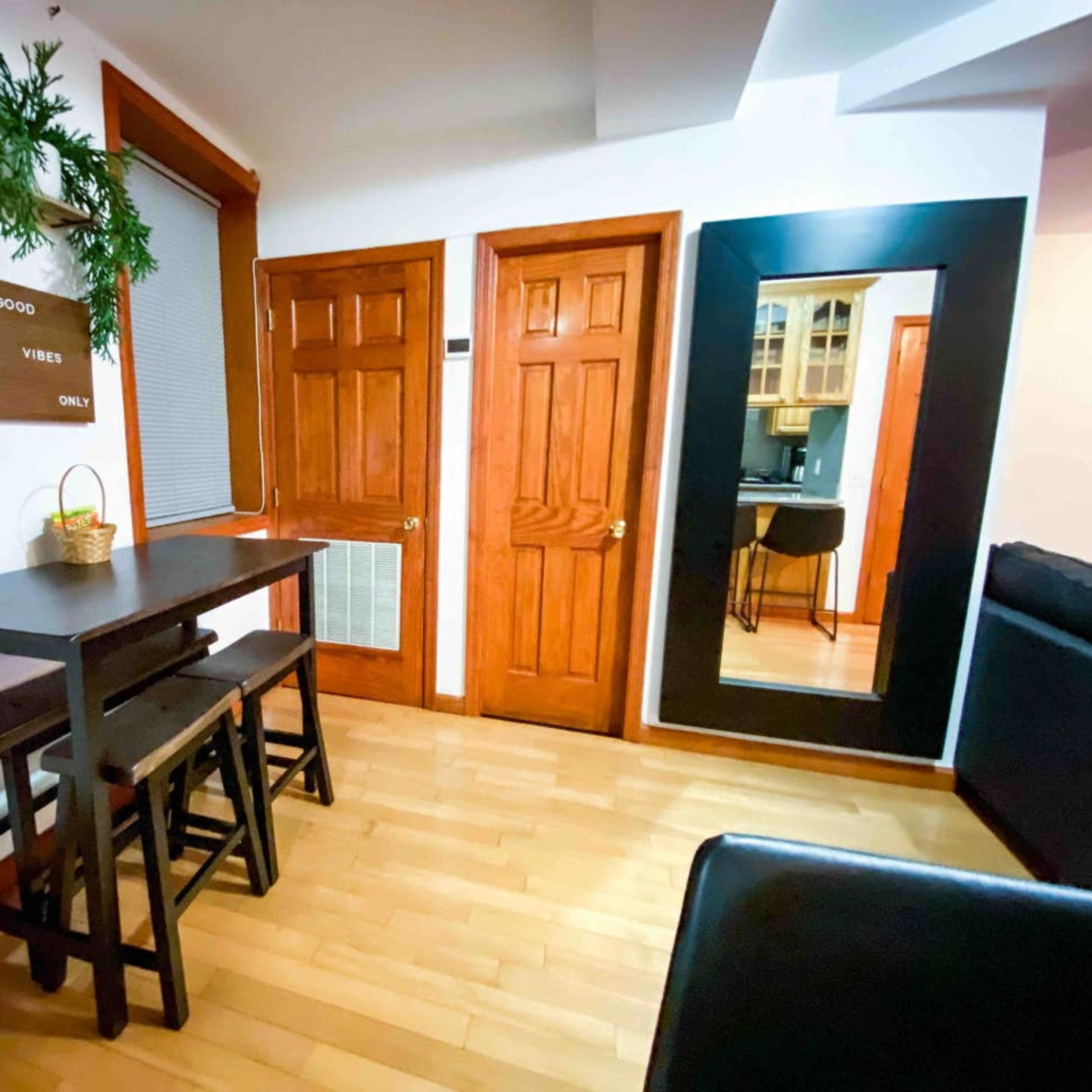 A wooden table with two stools, a wall mirror, and a decorative sign mounted on the wall in a room with wooden accents and natural light.